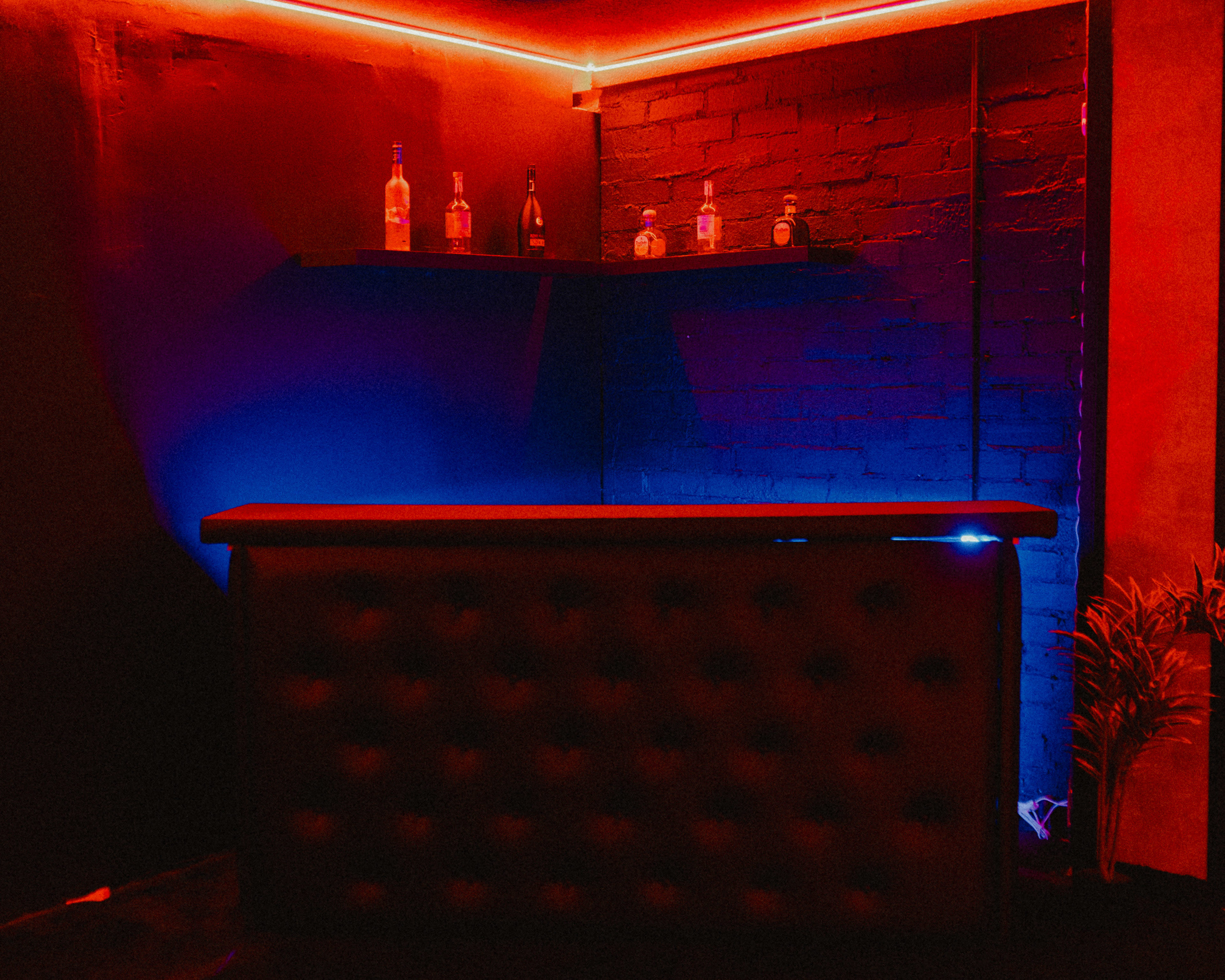 A bar with a backlit shelf holds several bottles against a wall illuminated by red and blue lights.