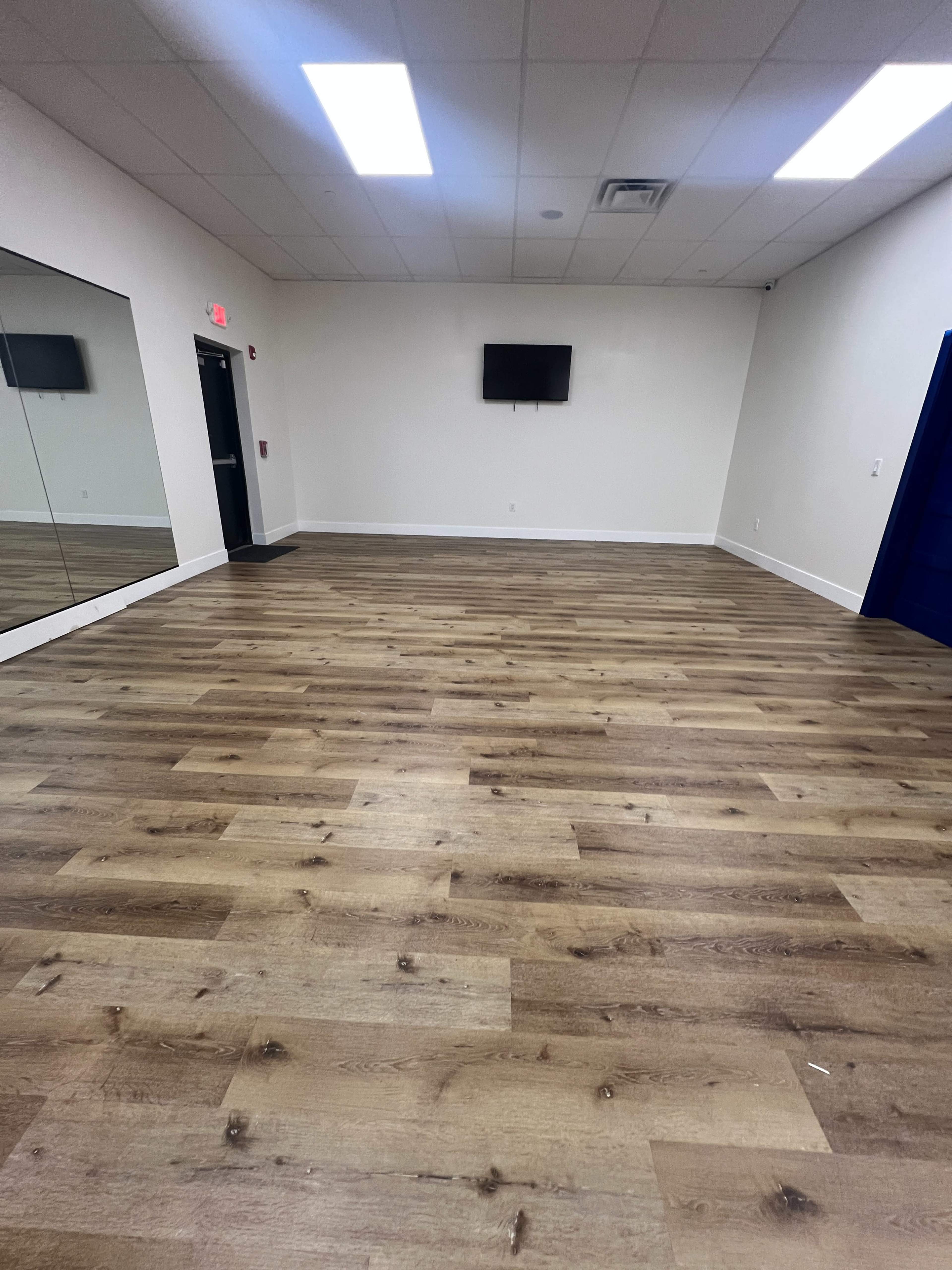 The image shows an empty room with wooden flooring, a large mirror on one wall, and a small television mounted on the opposite wall.