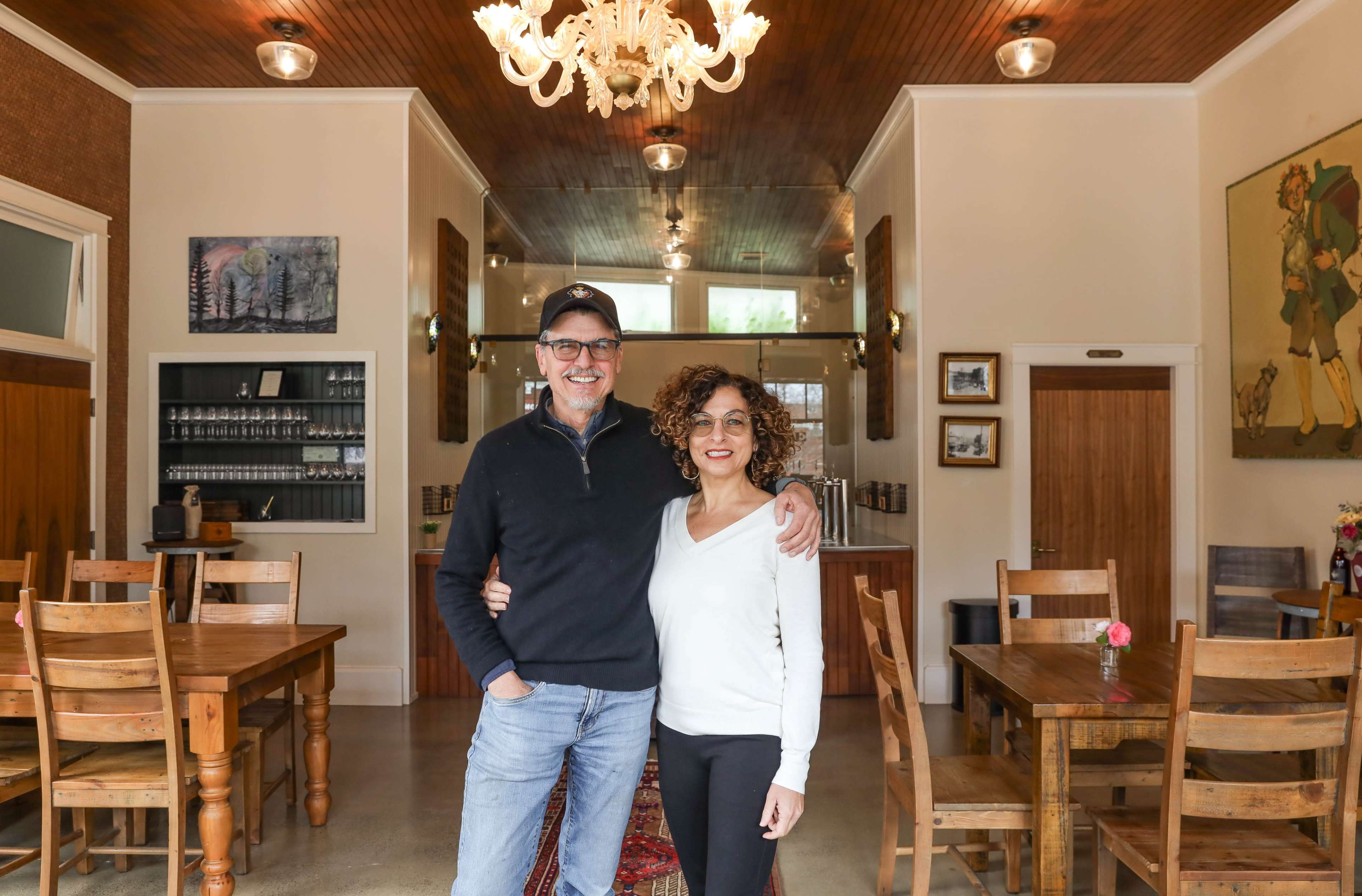 A man and a woman stand together smiling in a cozy café with wooden furniture and warm lighting.