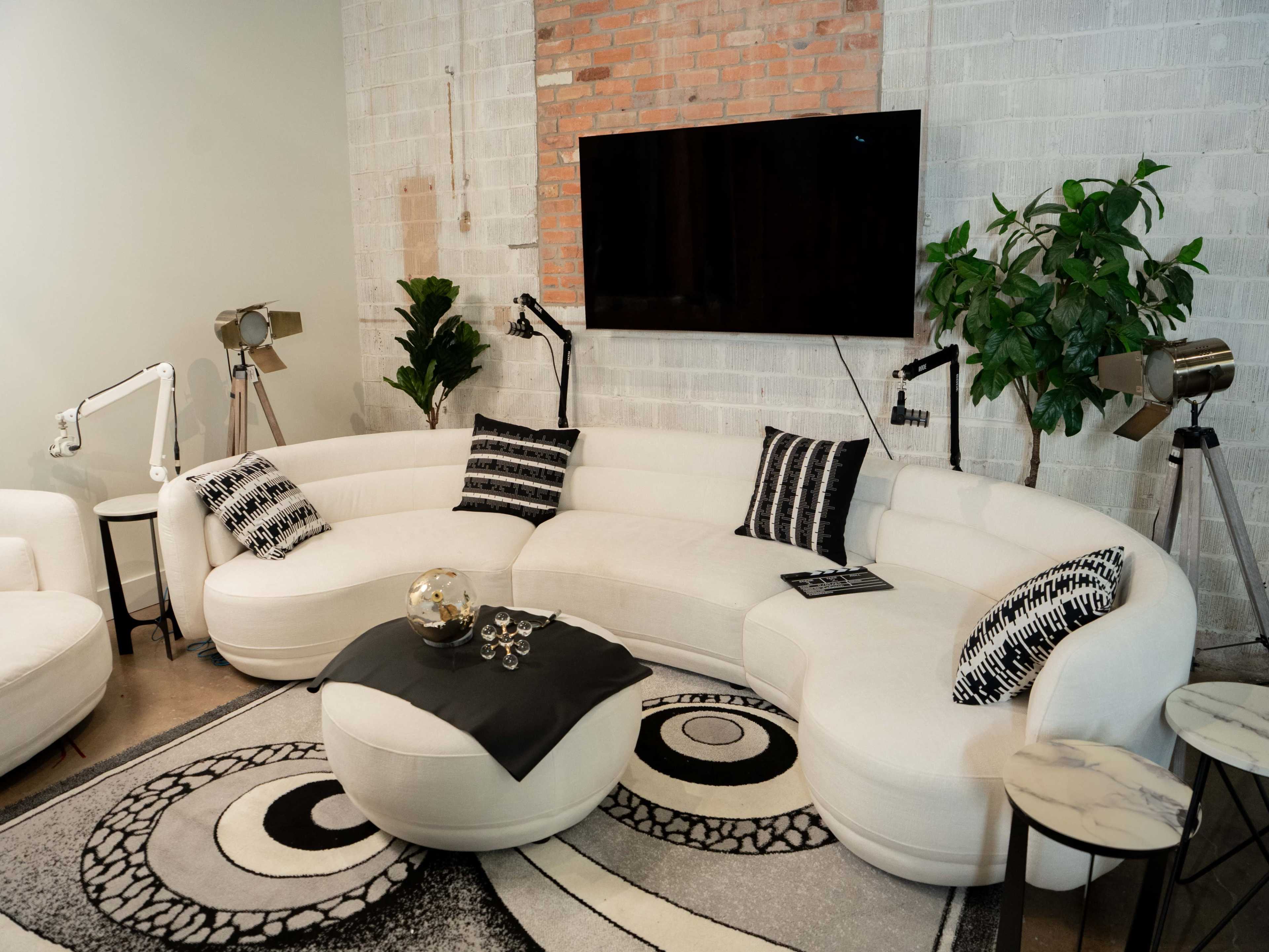 The image features a modern living room with a curved white sofa, a round coffee table, and decorative plants, all set against a brick wall with a mounted television.