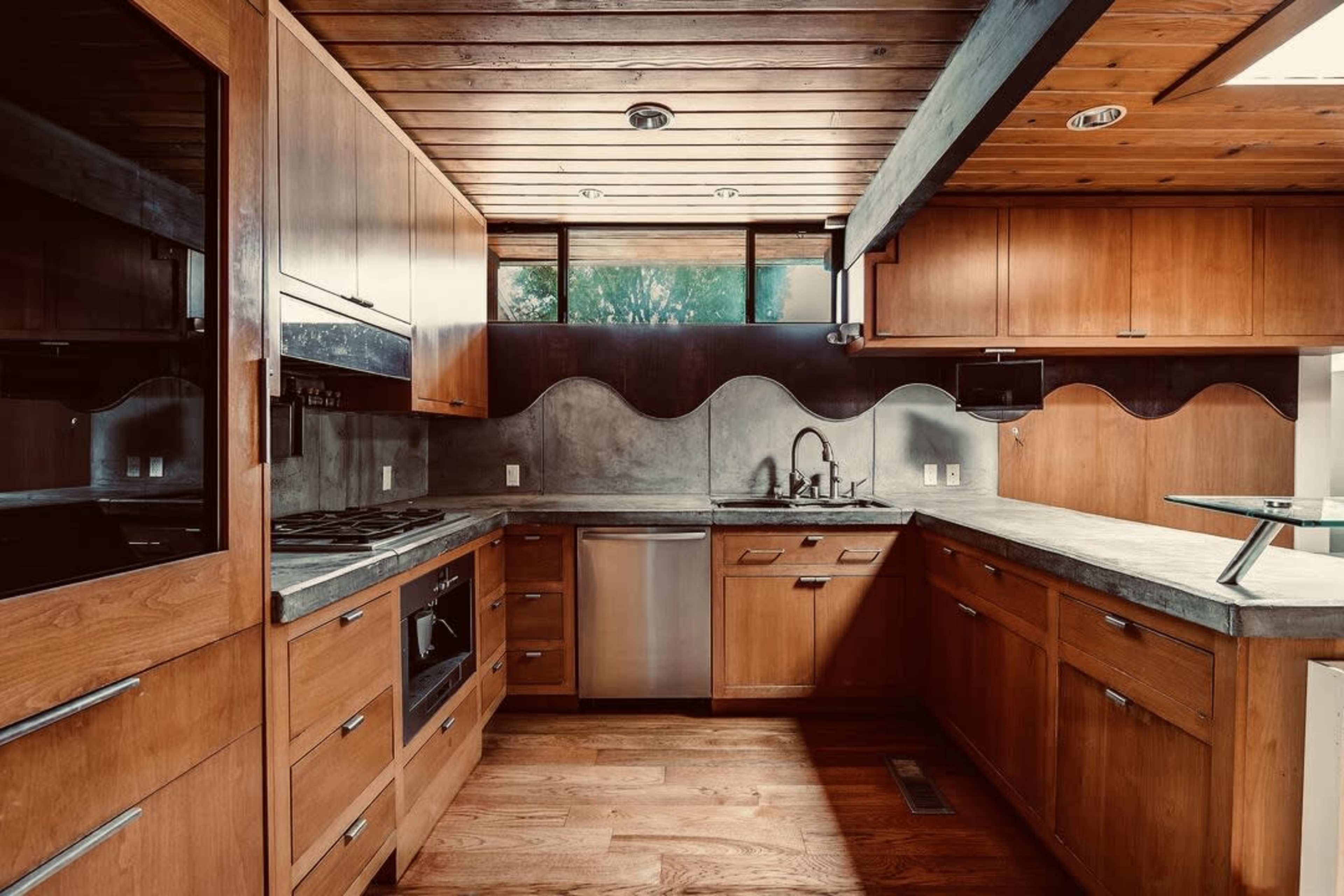 The image shows a modern kitchen featuring wooden cabinets, concrete countertops, and stainless steel appliances, with a large window providing natural light.
