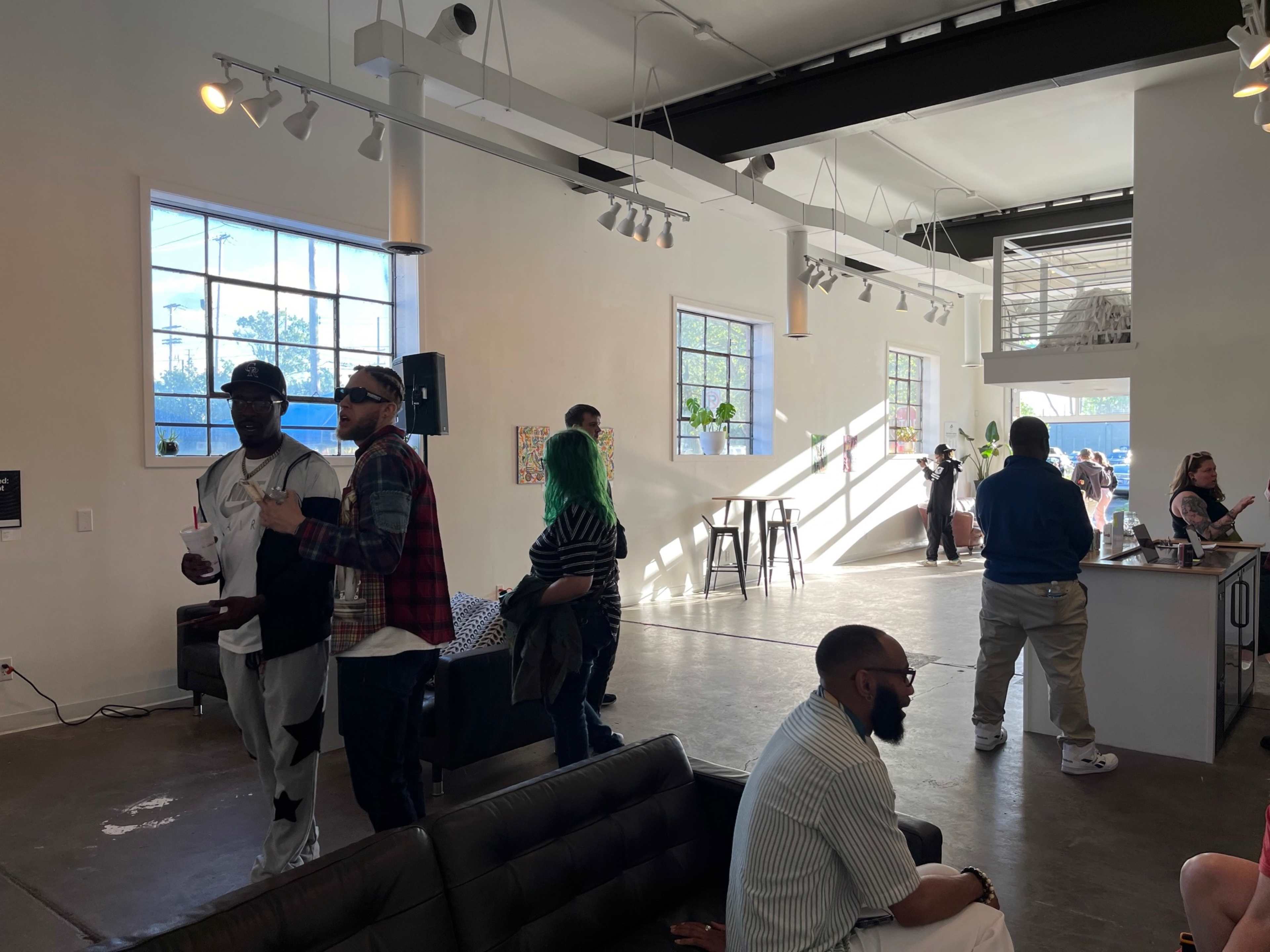 The image shows a group of people socializing in a spacious, well-lit art gallery with modern decor and large windows.