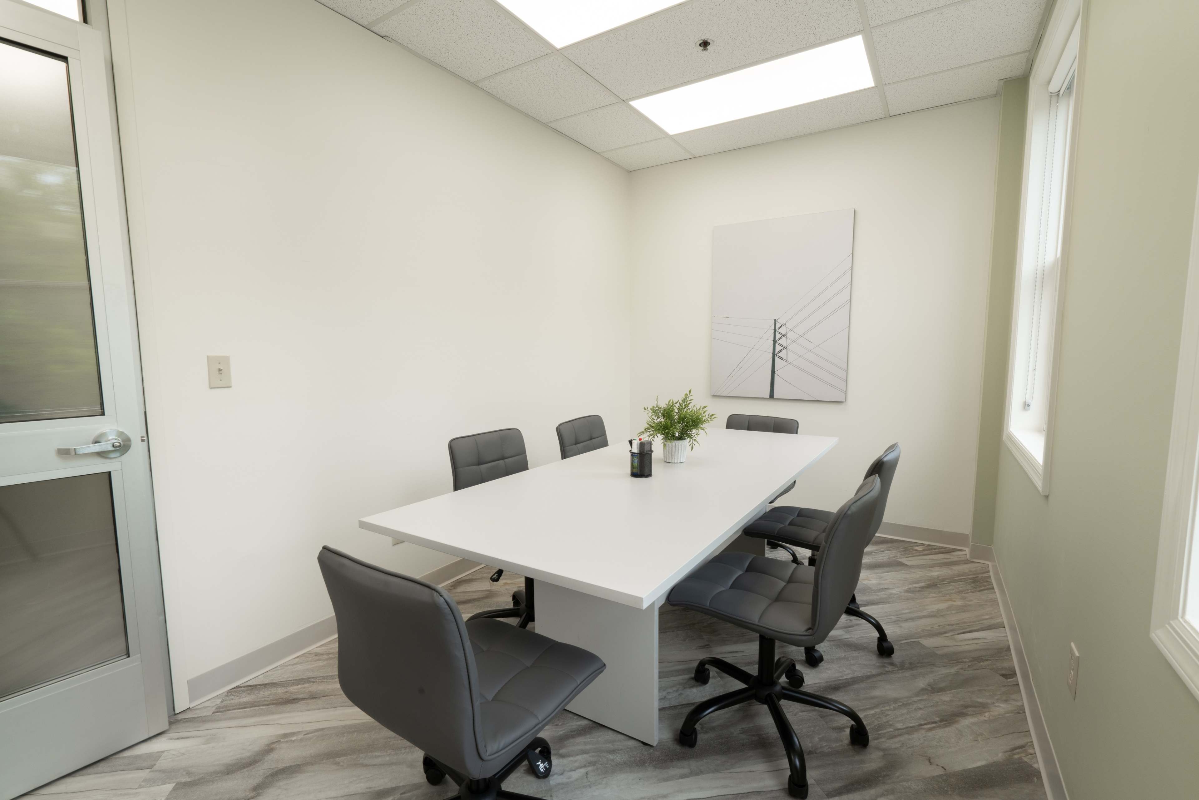 A minimalistic conference room features a central white table surrounded by four gray chairs, with a modern artwork on the wall.