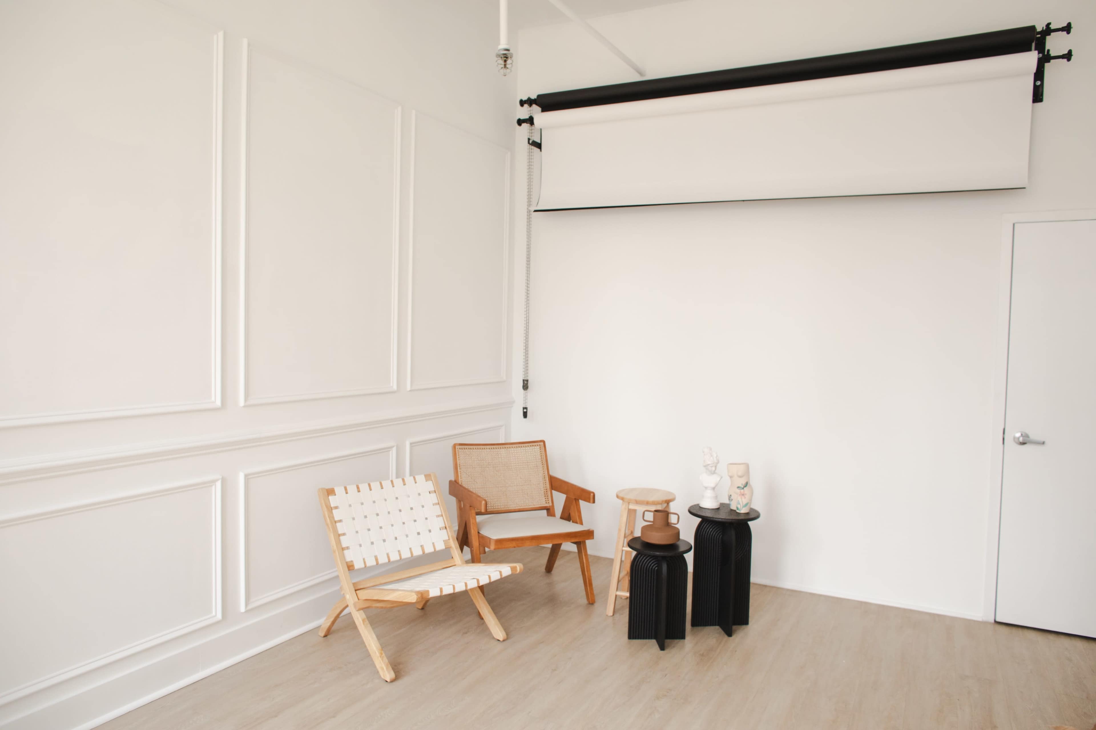 A minimalist interior room with two wooden chairs, a stool, and two small tables against a white wall.