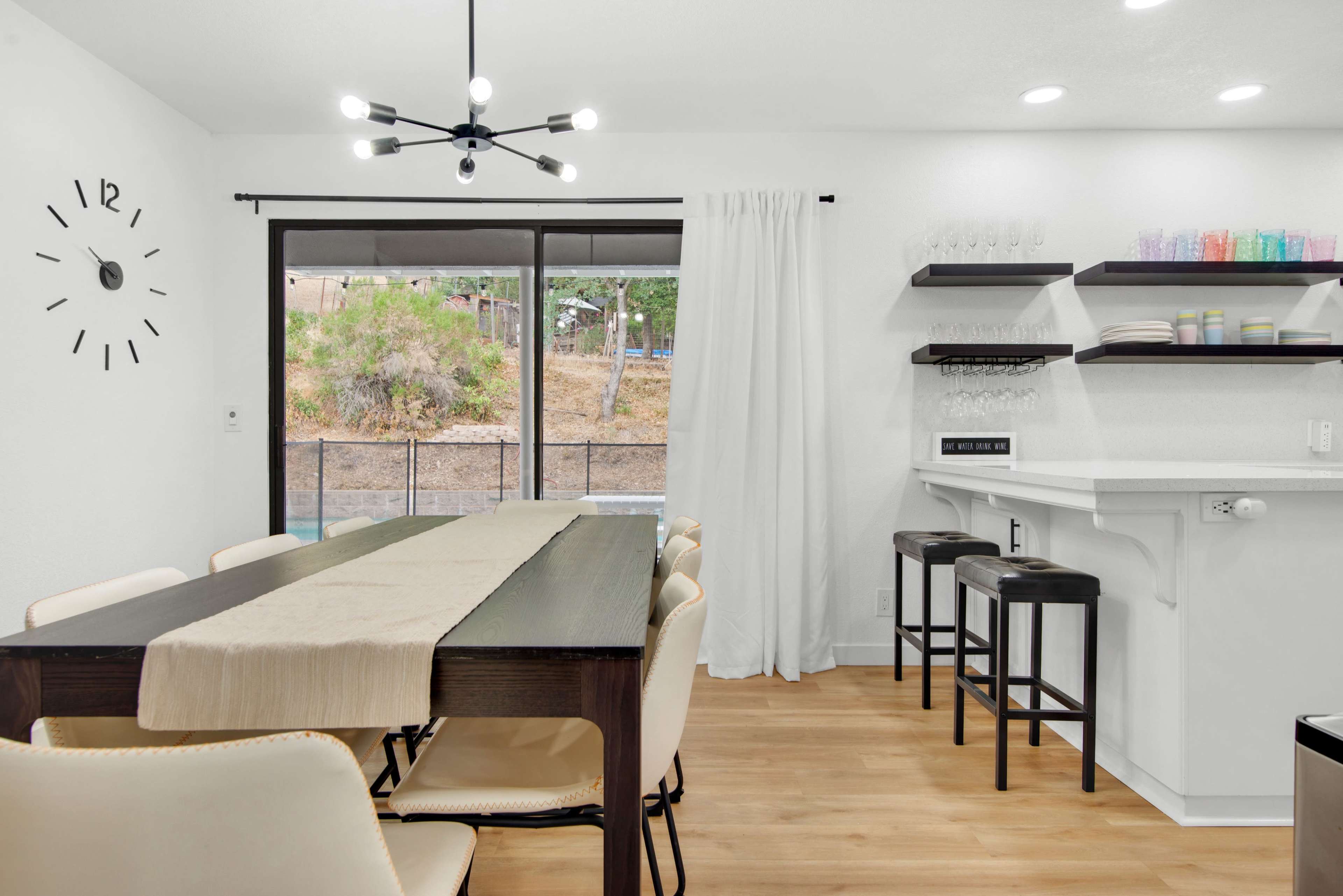 A modern dining area with a long table, chairs, and a bar counter, featuring a minimalist clock on the wall and large windows overlooking a garden.