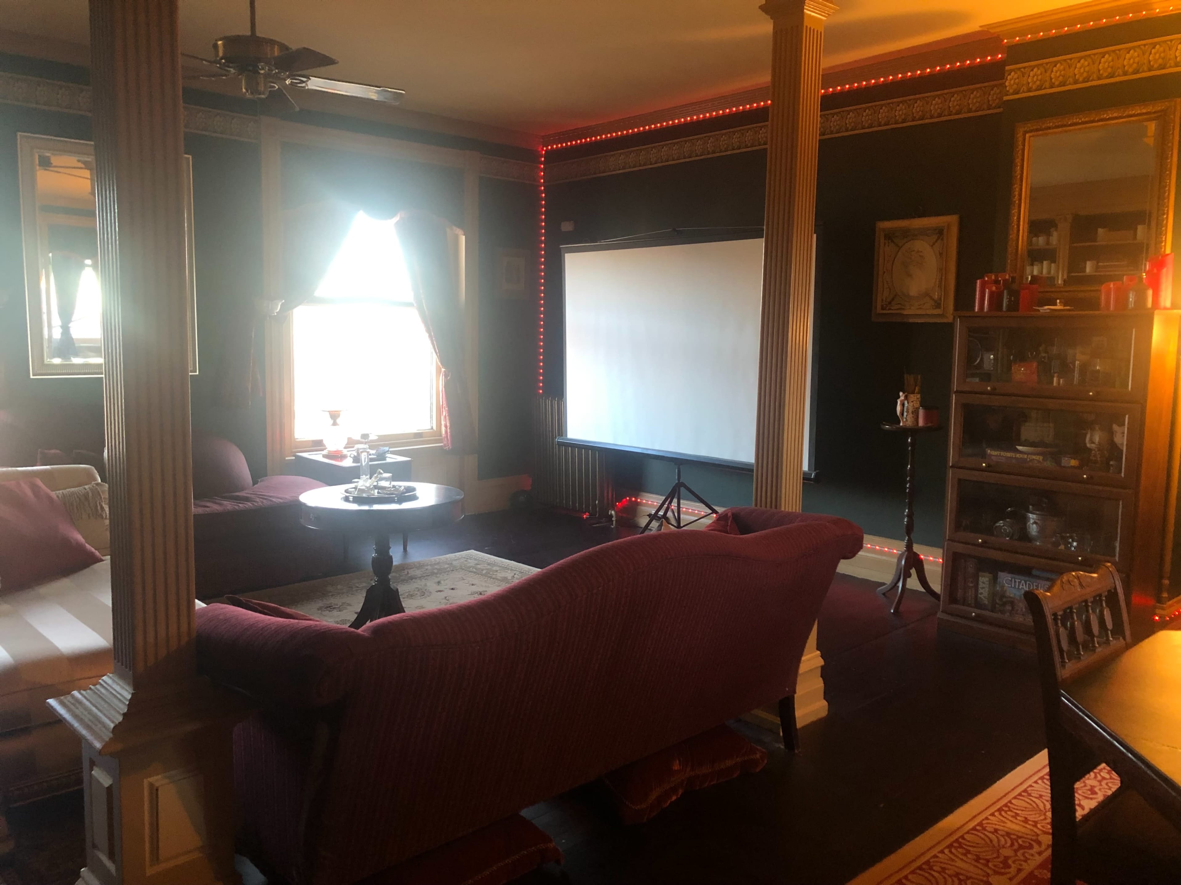 A living room features a large window with drapes, a screen for presentations, and a seating area with a maroon couch and a wooden table.