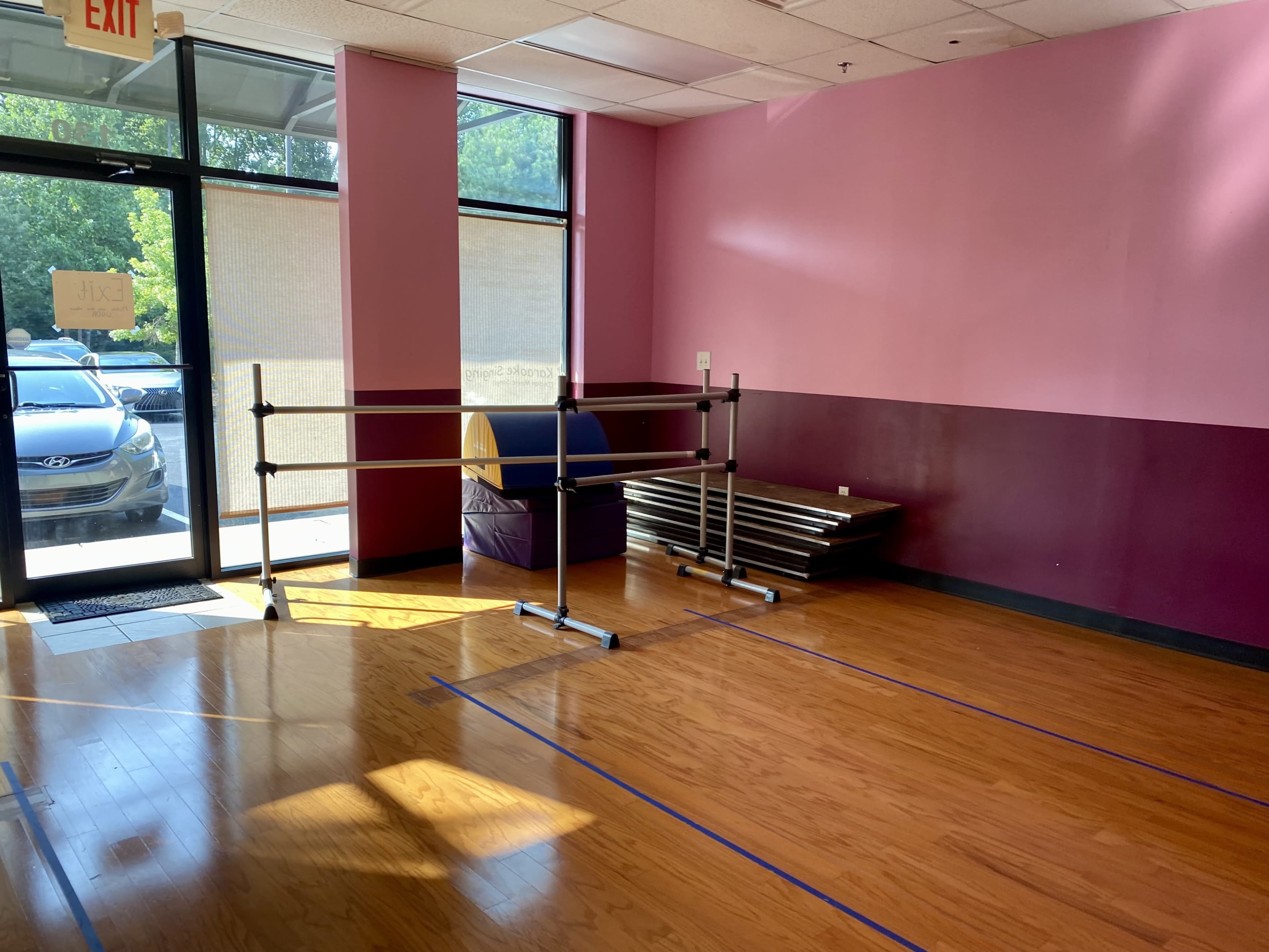 The room features a wooden floor, a ballet barre, and a purple mat, with large windows allowing natural light to enter.