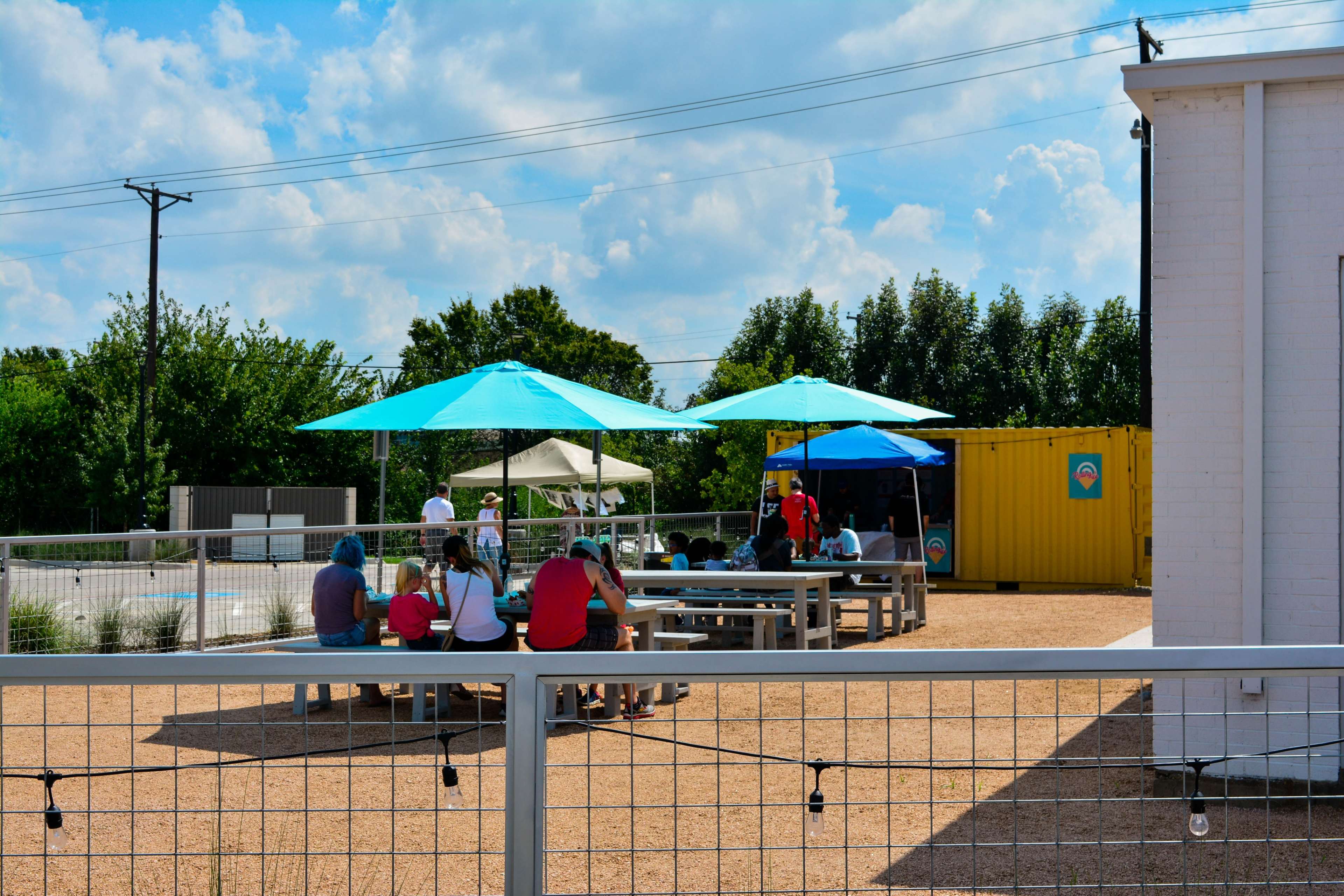 A group of people sits beneath umbrellas at outdoor tables in a casual outdoor gathering area near a yellow shipping container.