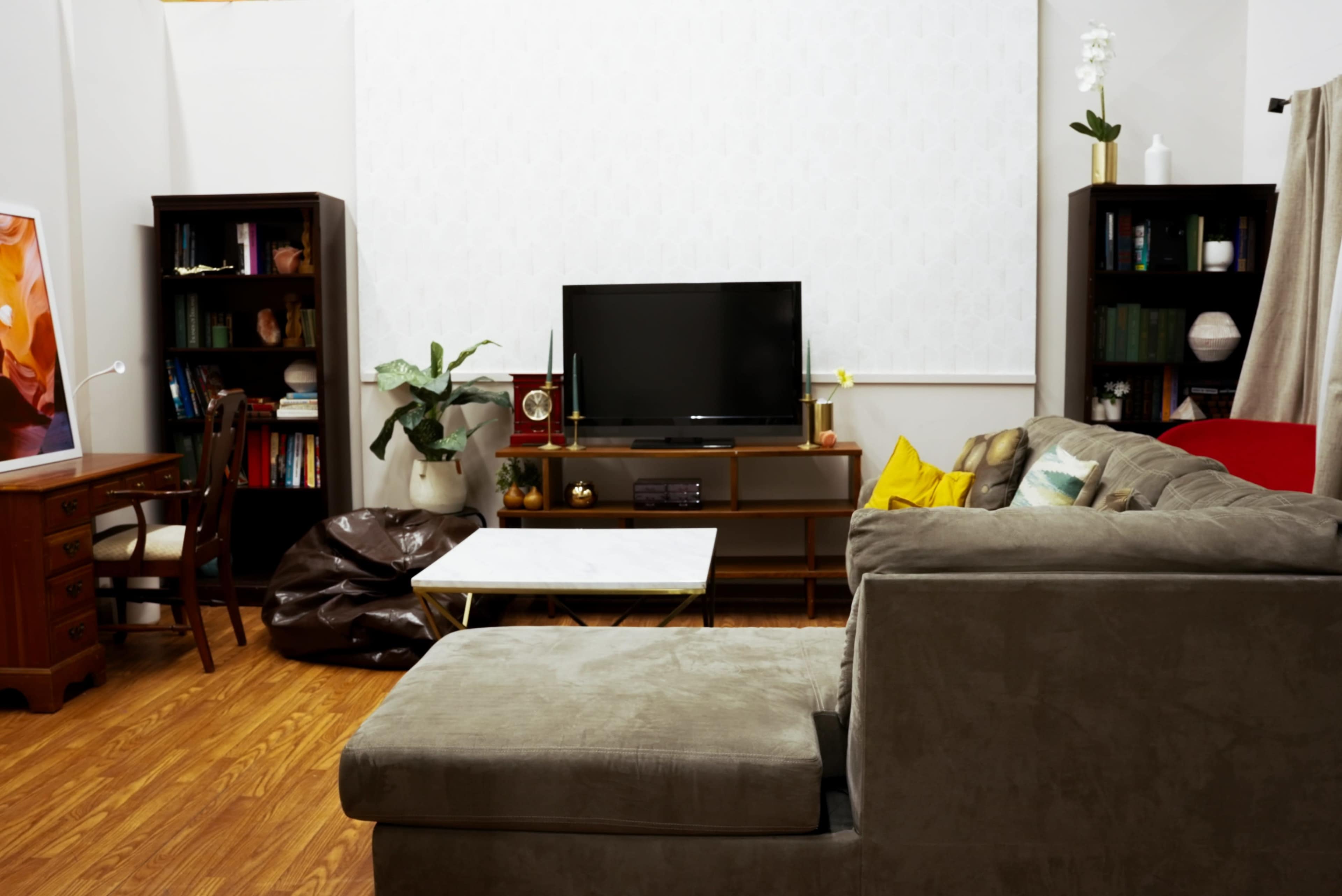 The image shows a modern living room with a television set, a wooden coffee table, and a sectional sofa, accompanied by bookshelves and a potted plant.