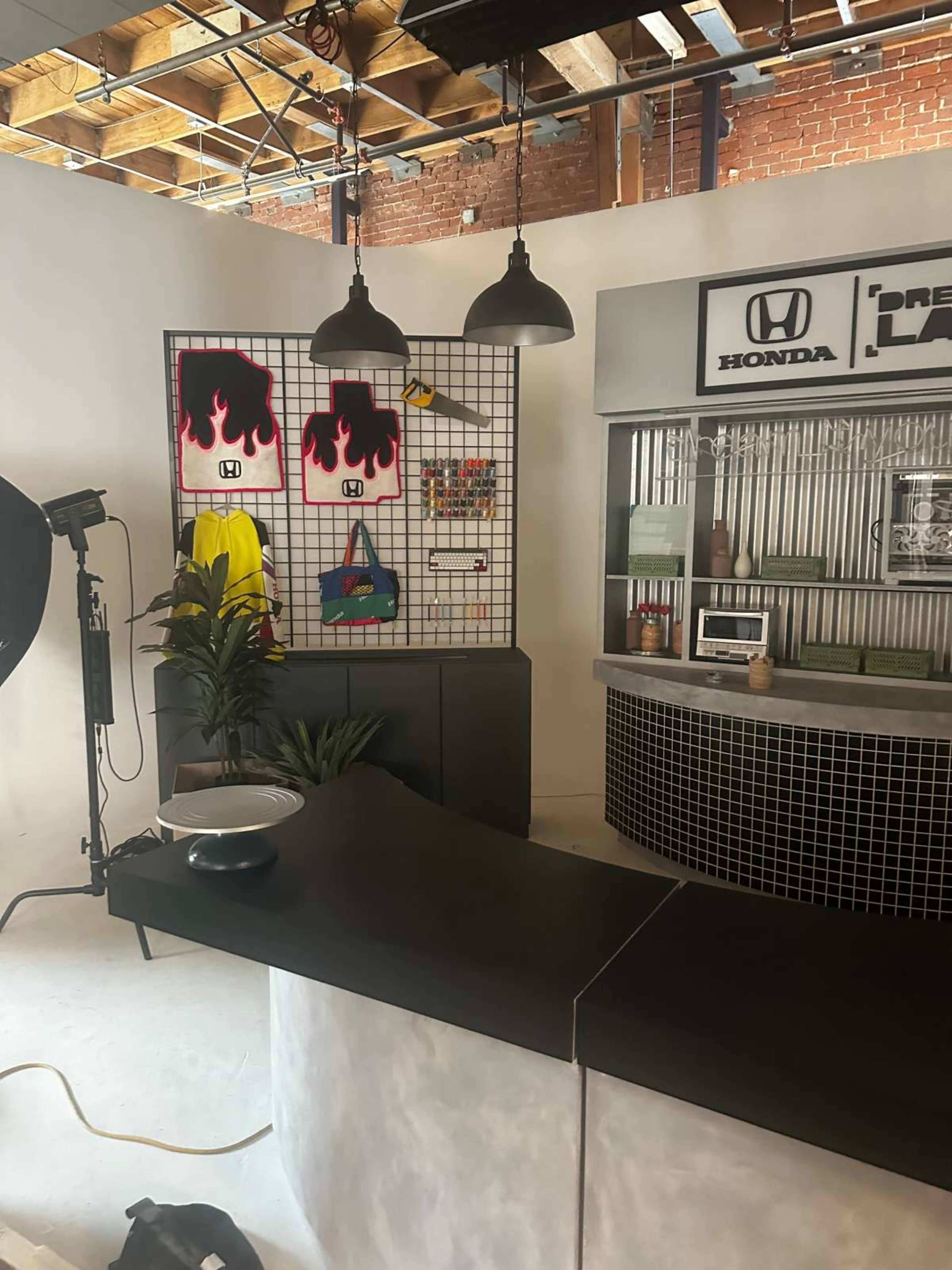 The image shows a modern studio space featuring a curved black countertop, a grid display with colorful items, and a Honda-branded area with plants and industrial lighting.
