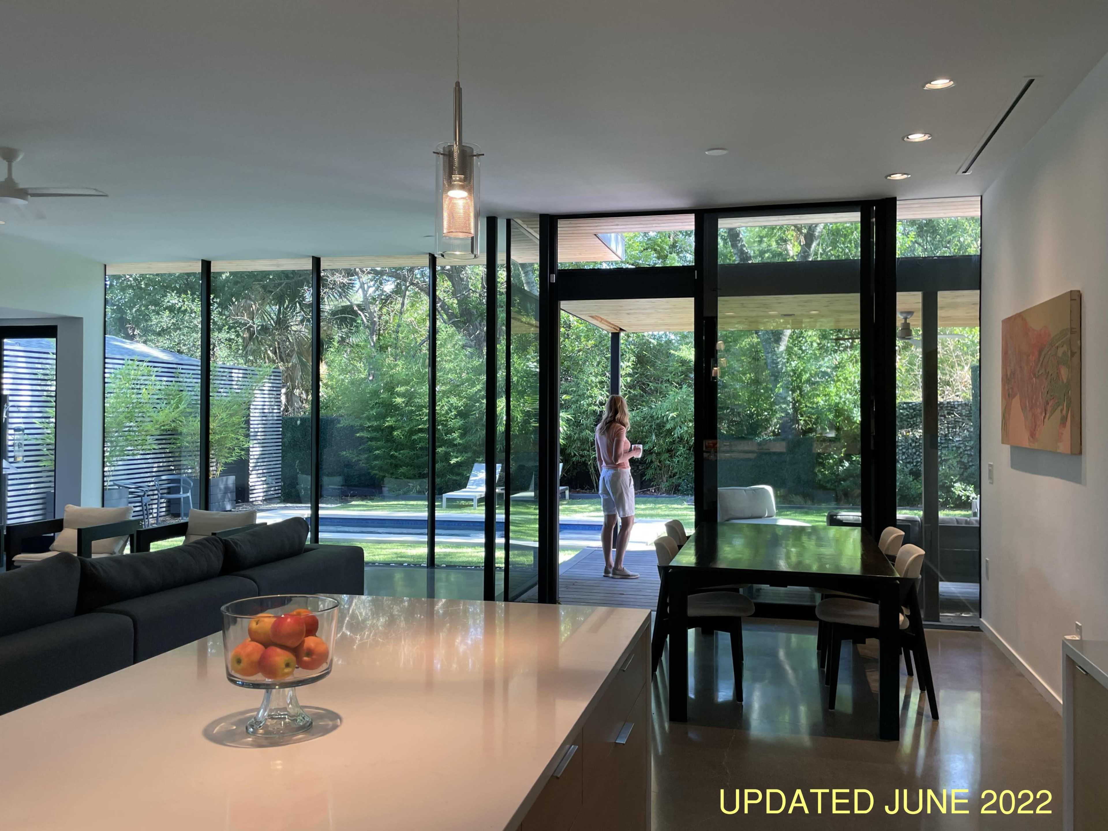 A woman stands near large glass windows overlooking a garden from a modern kitchen with a fruit bowl on the counter.