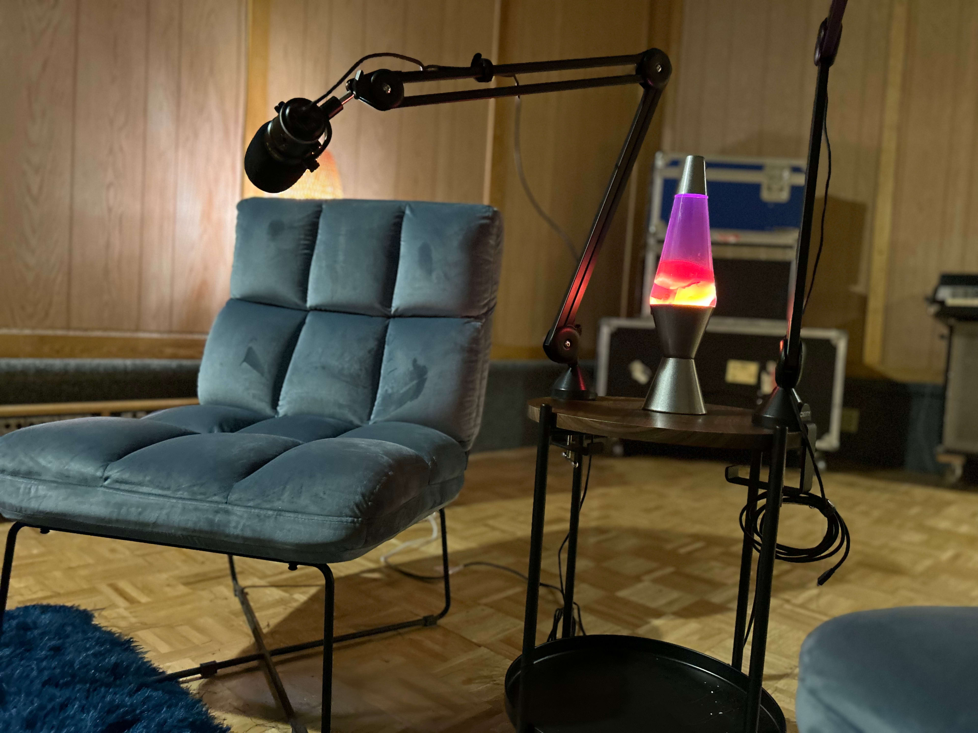A blue velvet chair with a microphone attached to an adjustable arm sits next to a table featuring a lit lava lamp in a room with wooden paneling.