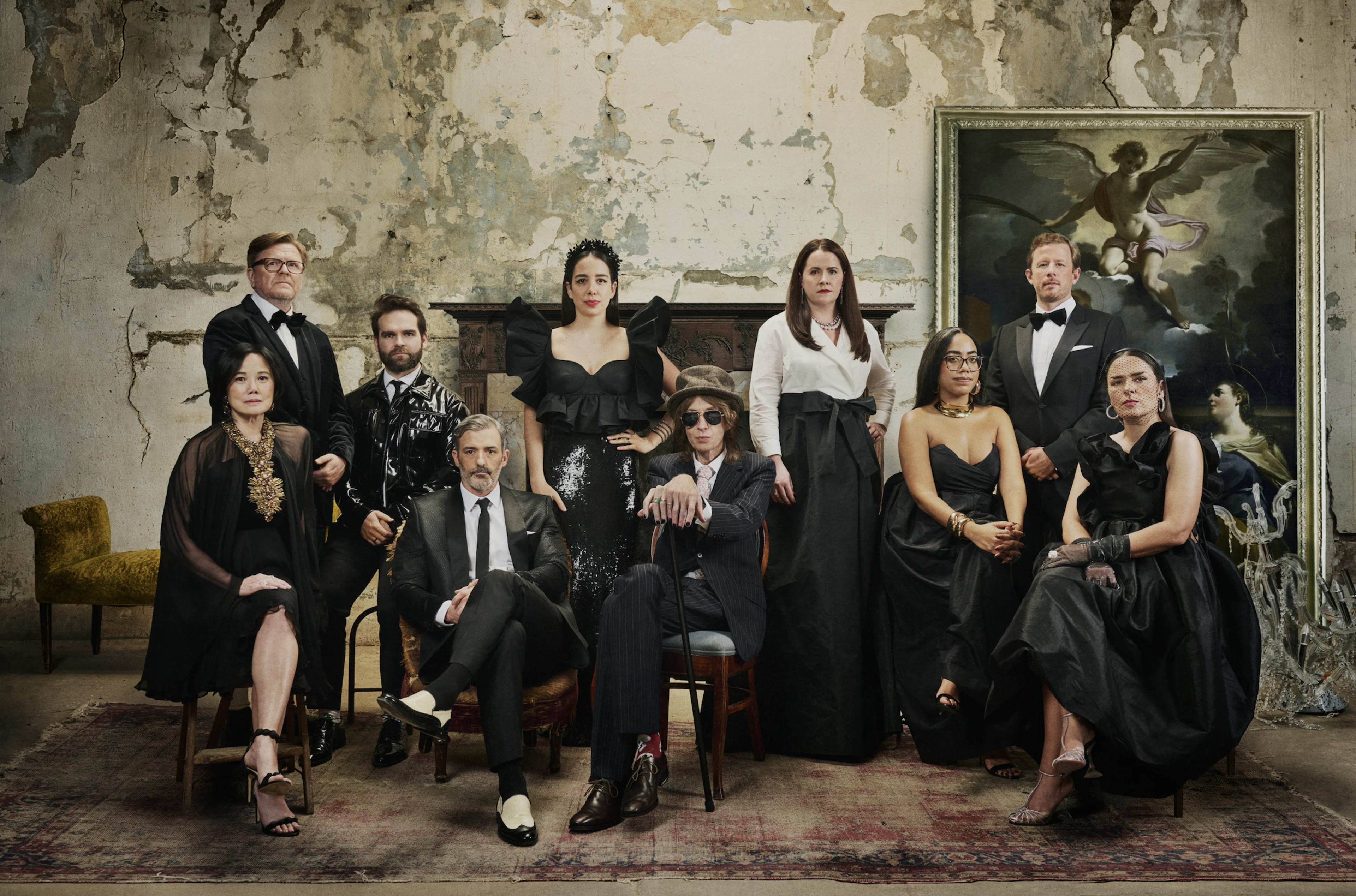A group of twelve individuals dressed in formal black attire pose together in an ornate but decaying room with a large painting on the wall.