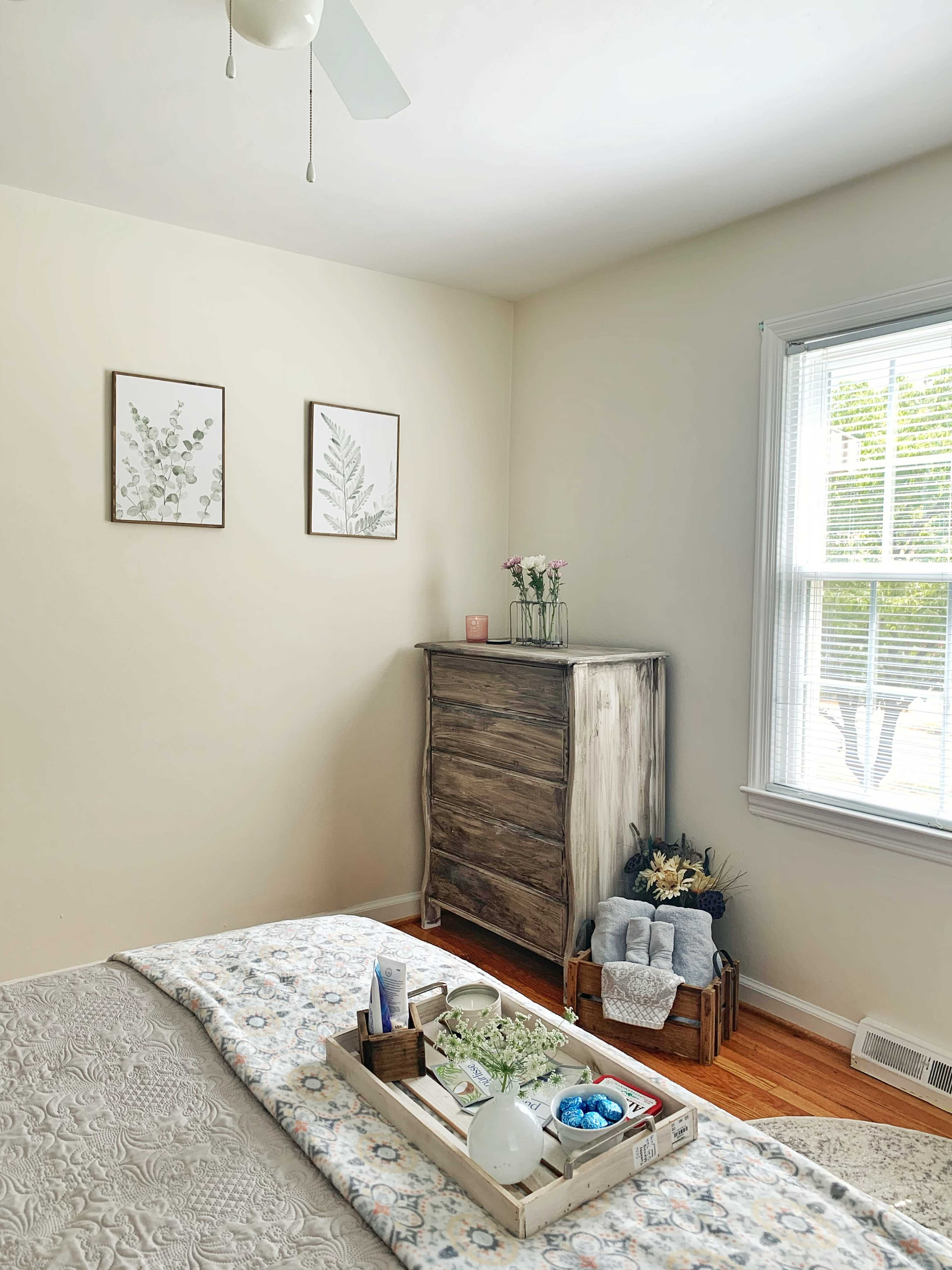 A neatly arranged bedroom features a bed with a decorative throw, a wooden dresser, and framed botanical prints on the wall near a window.