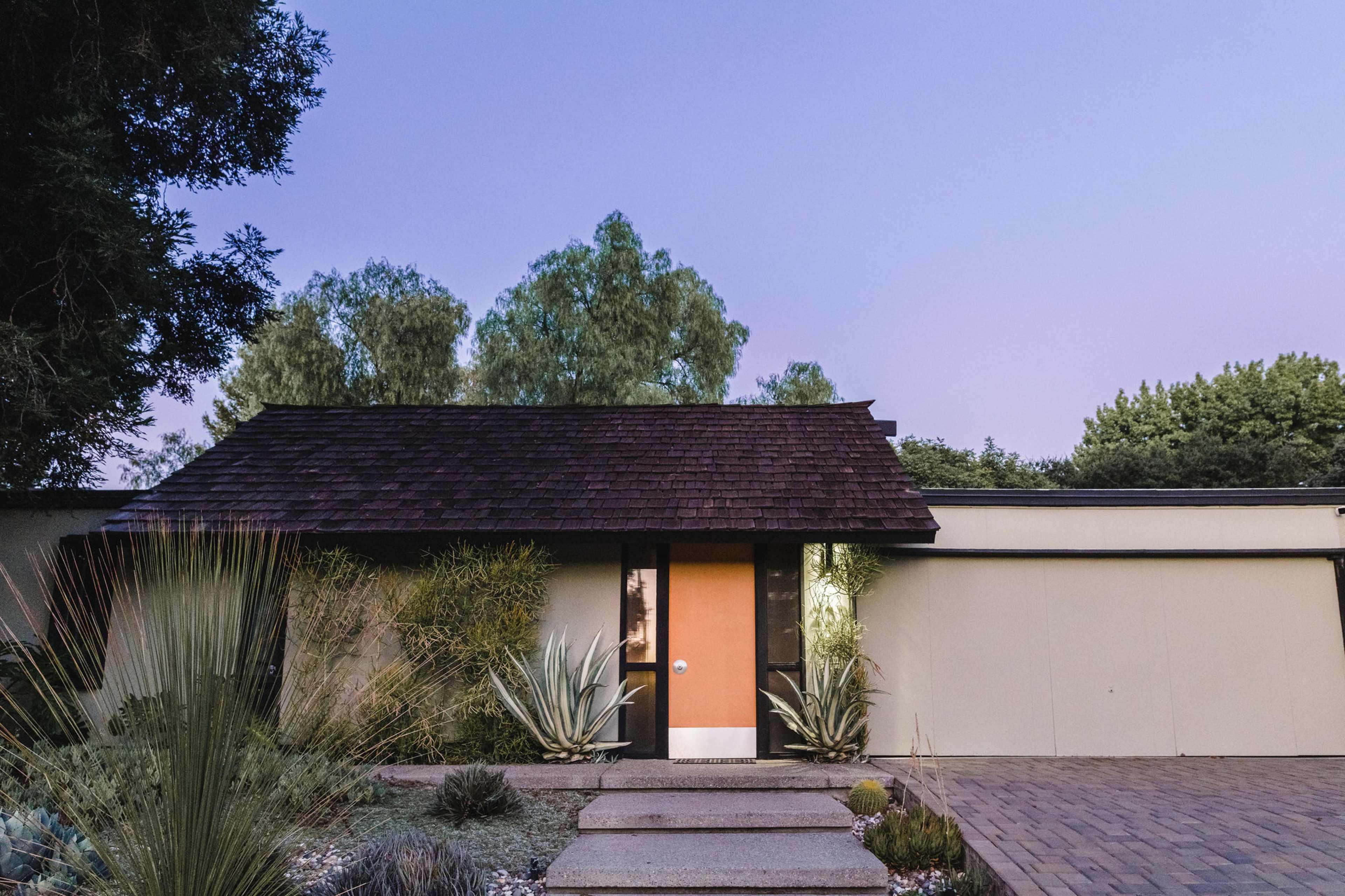 A modern house with a sloped roof and an orange door, surrounded by low landscaping and pathways.