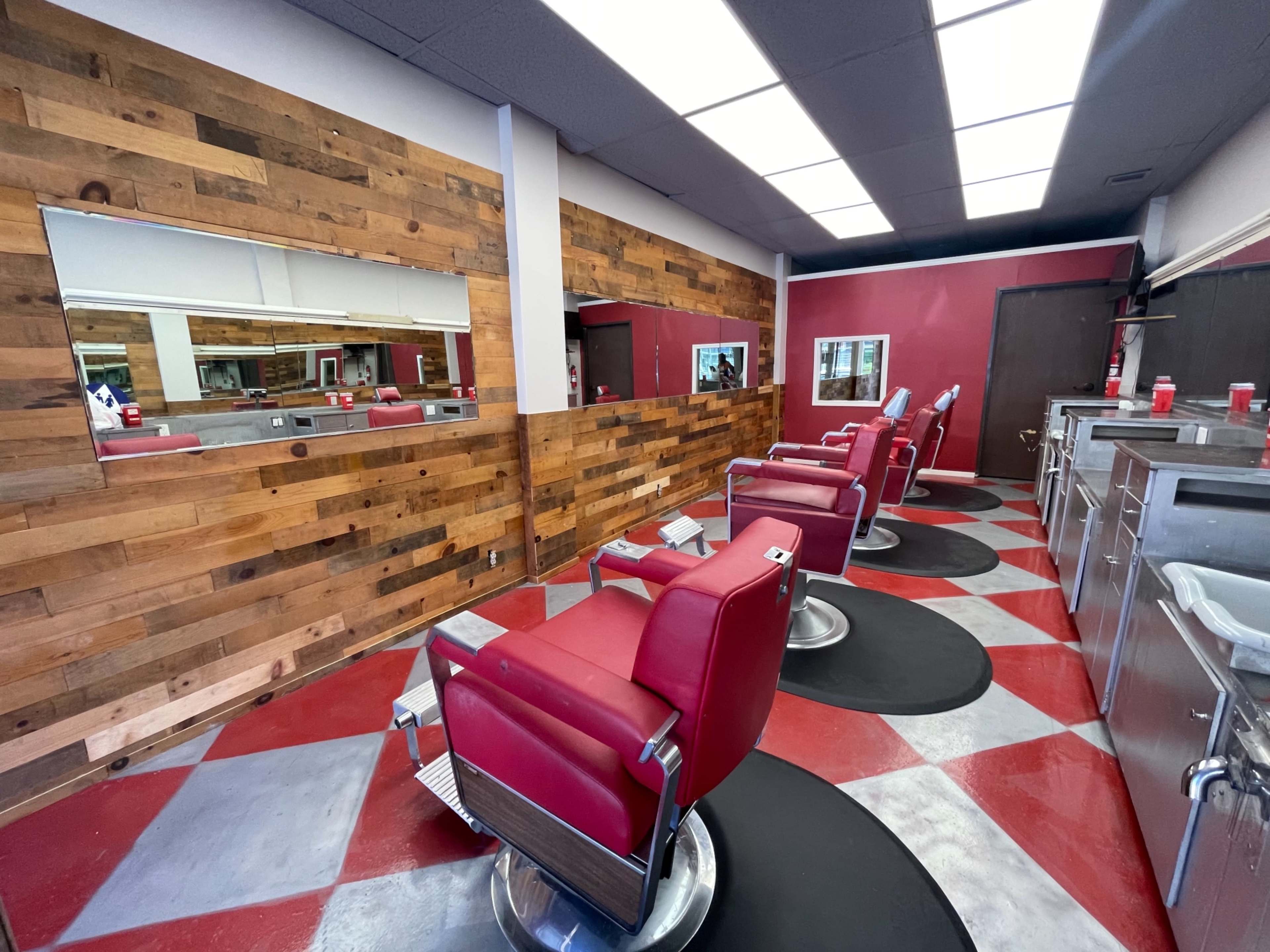 A barber shop interior with three red barber chairs, a wooden accent wall, and checkered flooring.