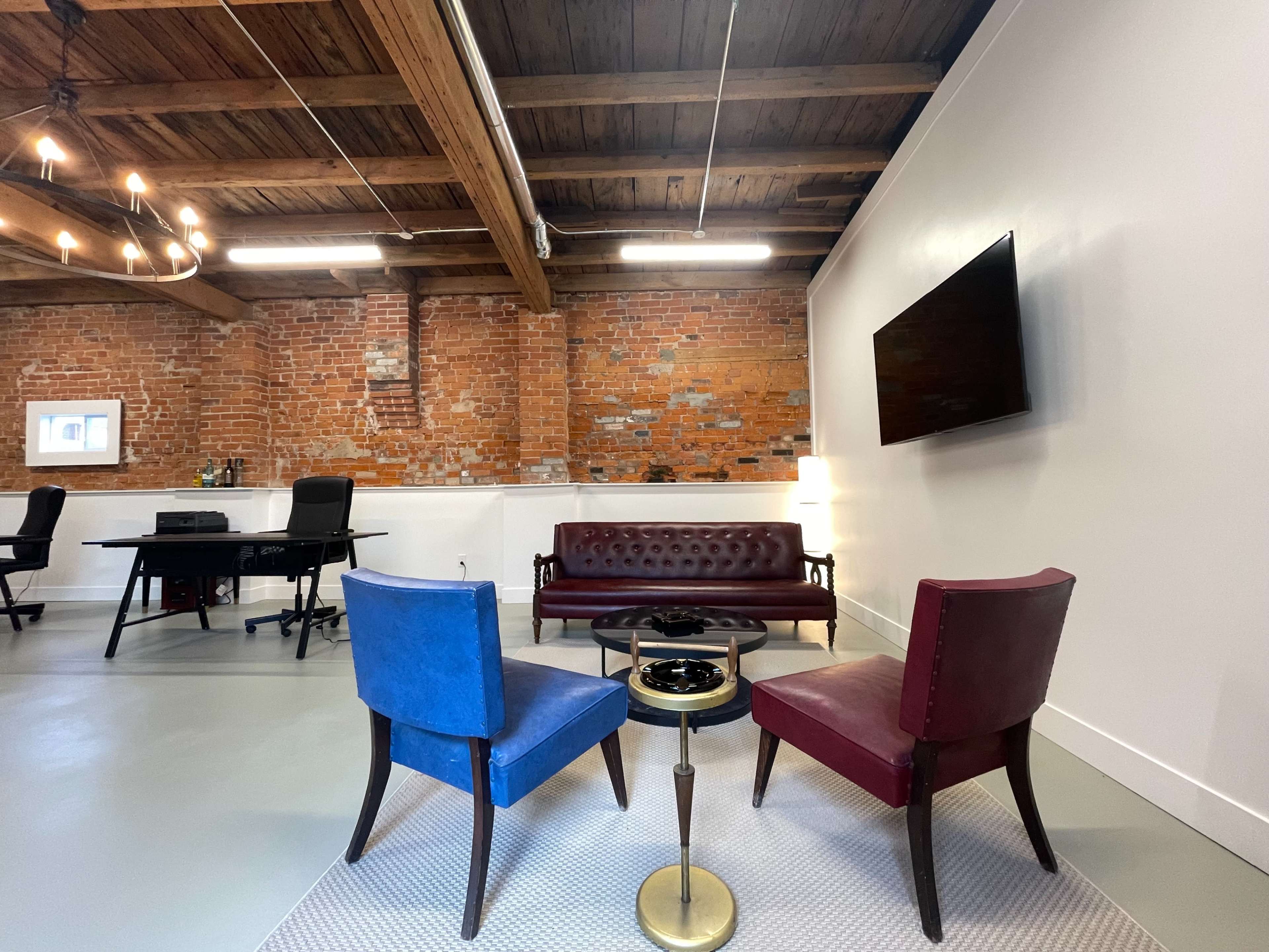A modern office interior with a brown leather sofa, two colorful chairs, a small table, and exposed brick walls.