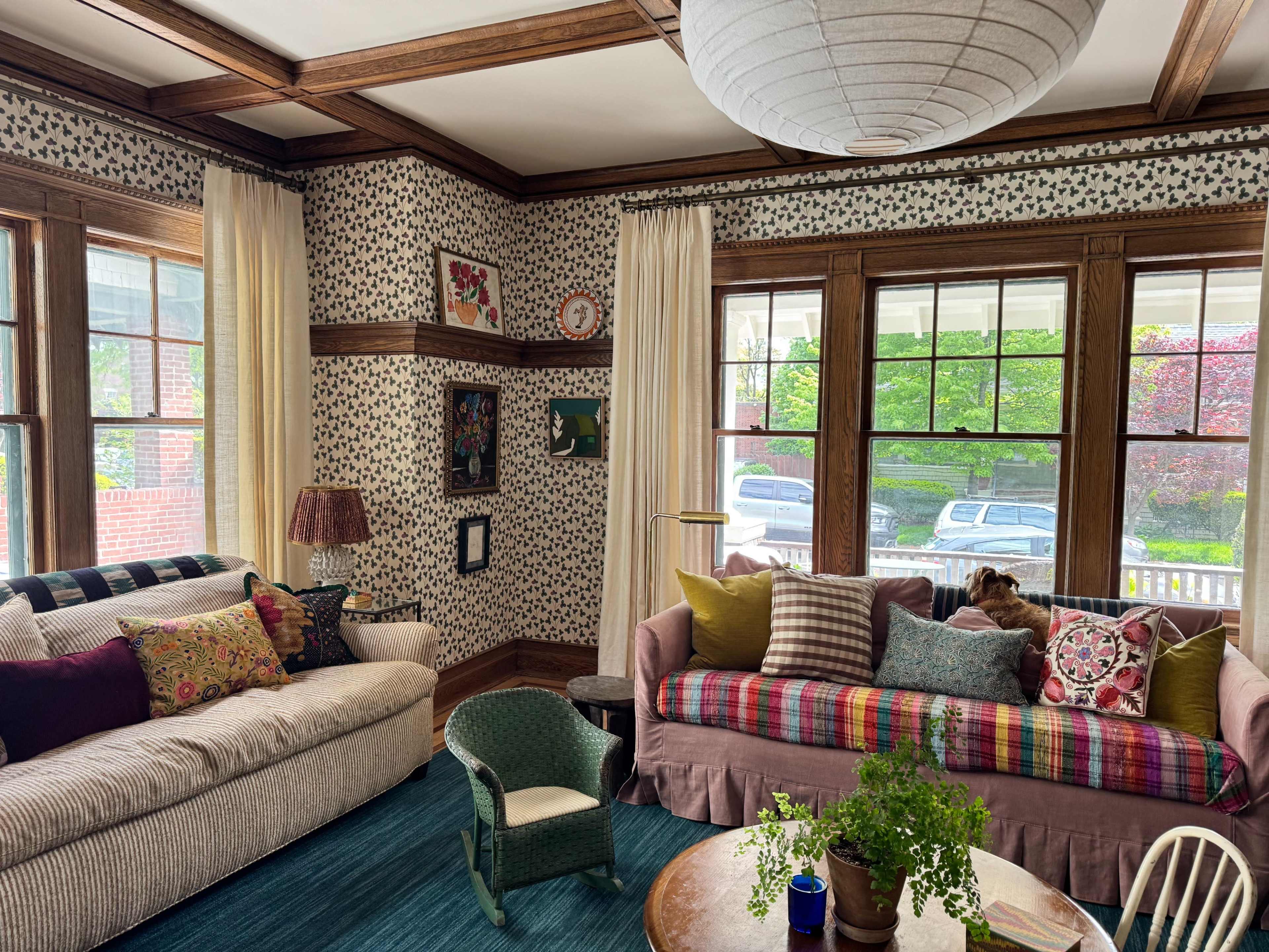 The image shows a cozy living room with patterned wallpaper, two couches adorned with various pillows, a round wooden table, and large windows letting in natural light.