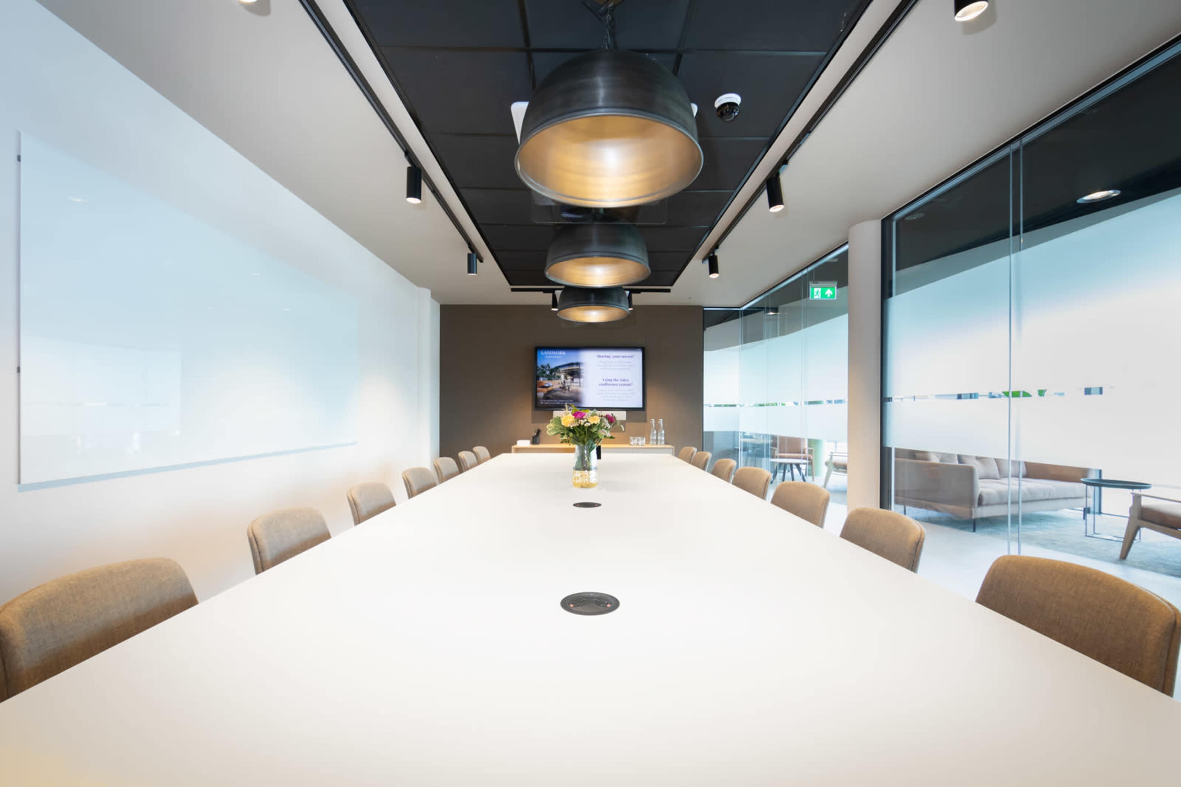 A modern conference room features a long white table surrounded by beige chairs, with pendant lighting overhead and a large screen at one end.