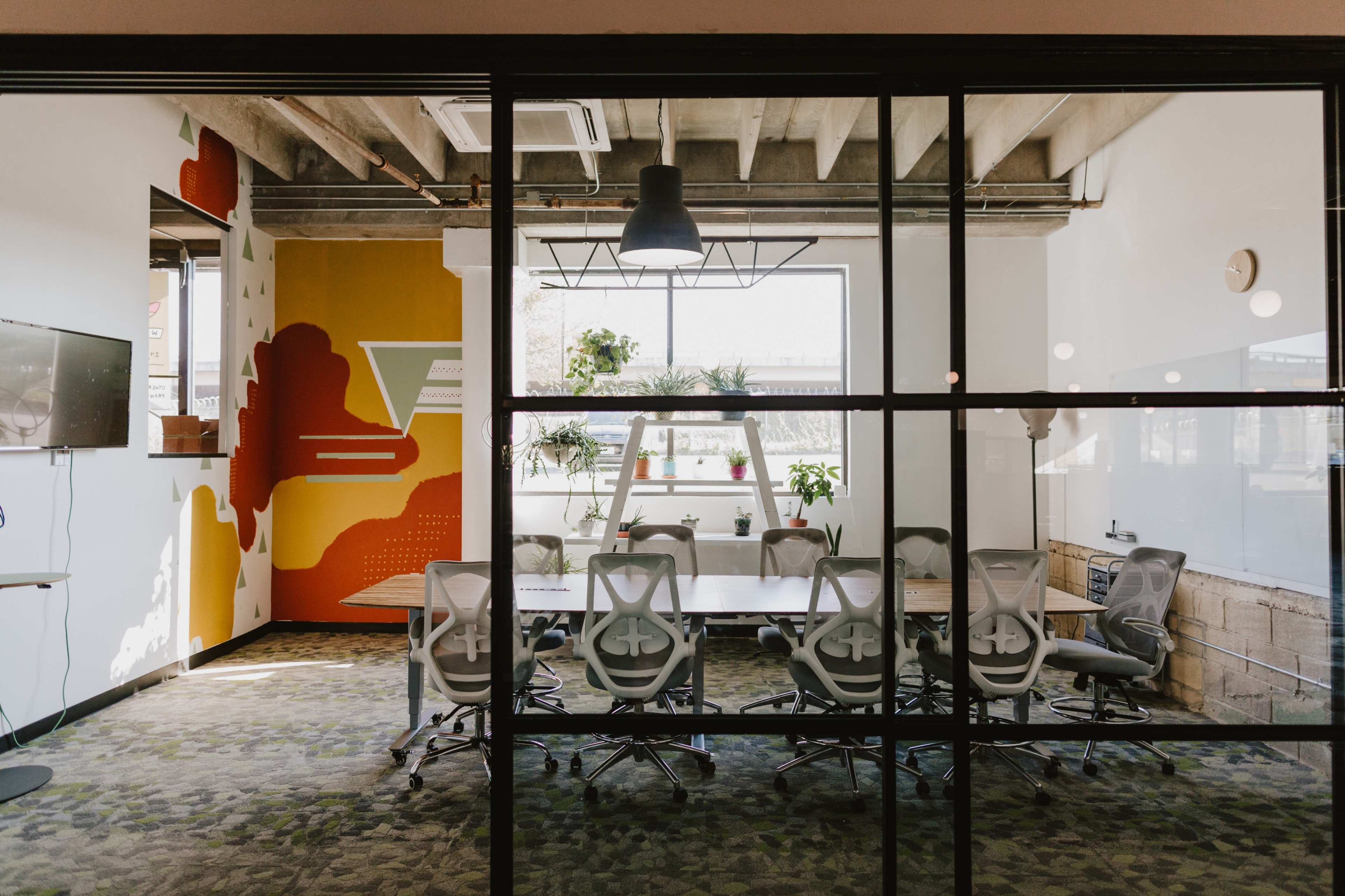 A modern conference room features a large table surrounded by office chairs, with colorful wall art and plants visible through glass walls.