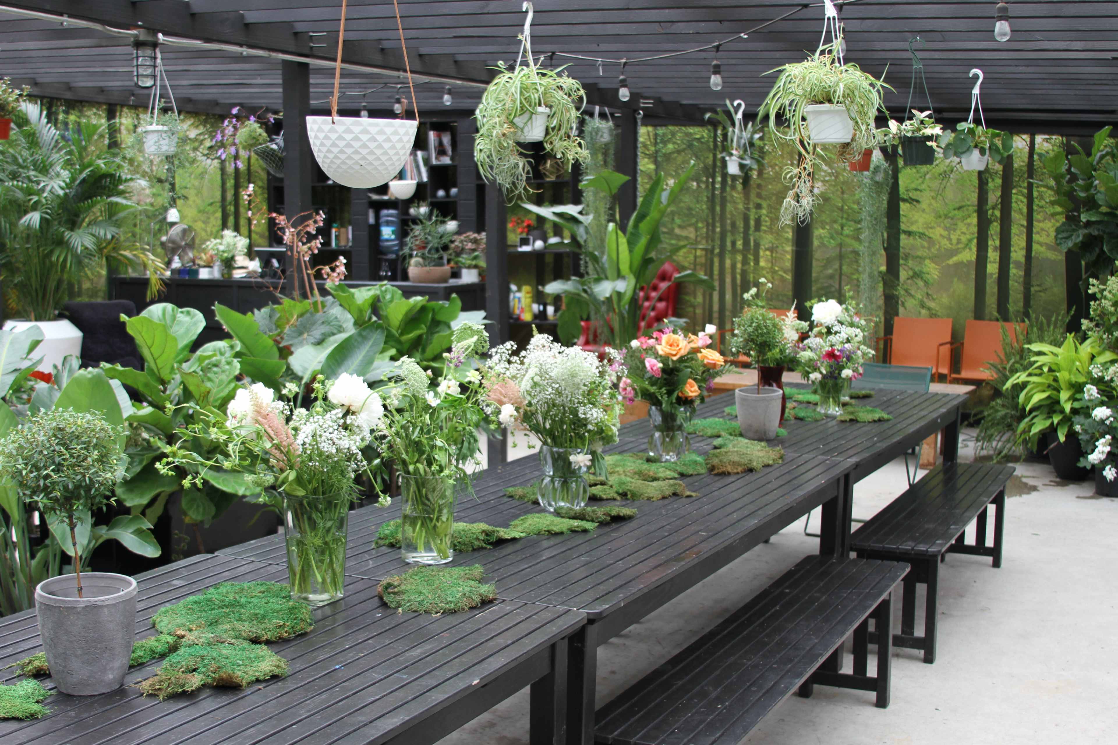 A long black wooden table is set under a shaded pergola, adorned with vases of flowers and surrounded by greenery and hanging plants.