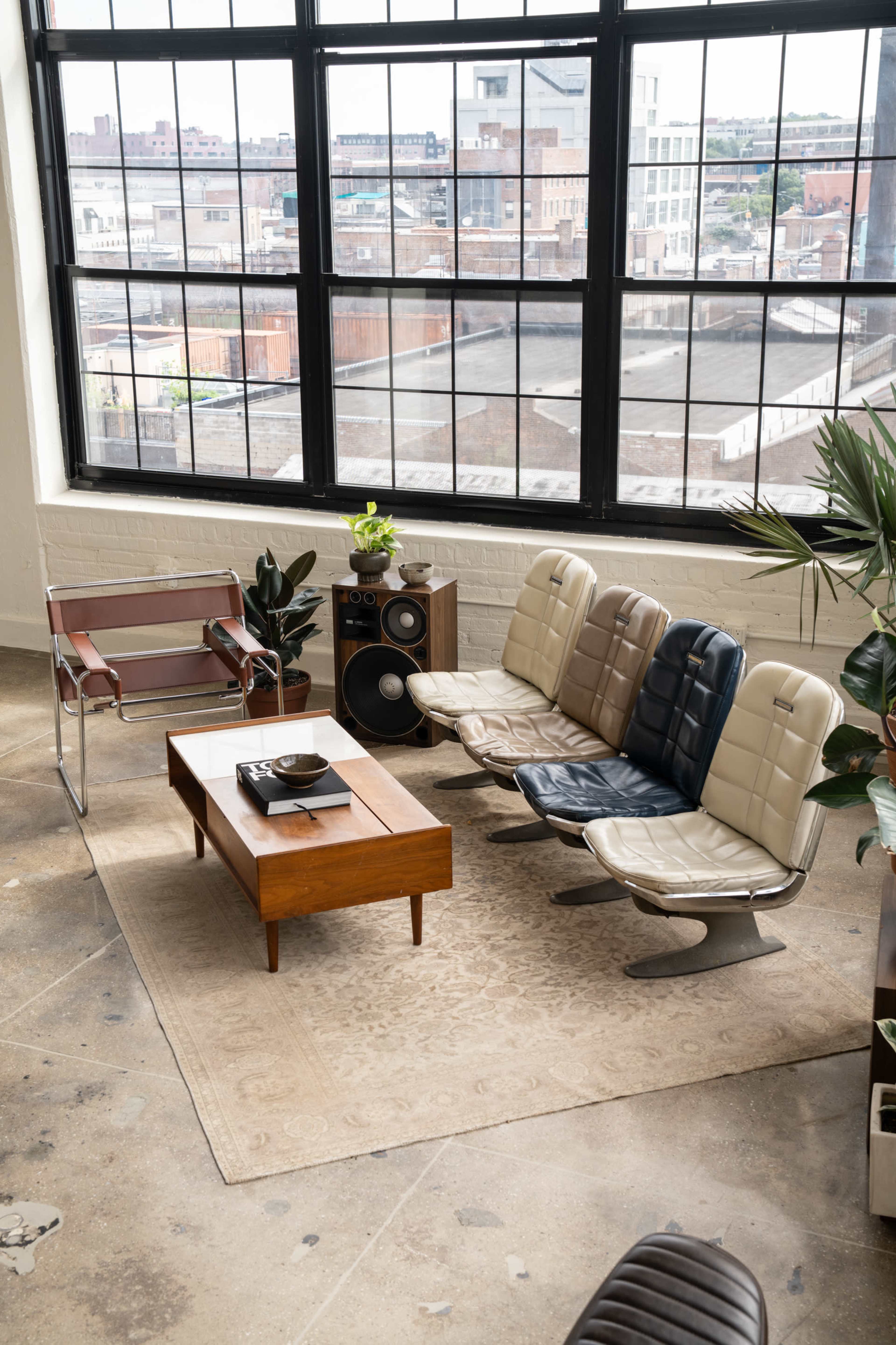 The image shows a stylish living room with modern chairs arranged around a wooden coffee table, large windows letting in natural light, and a decorative plant in the corner.