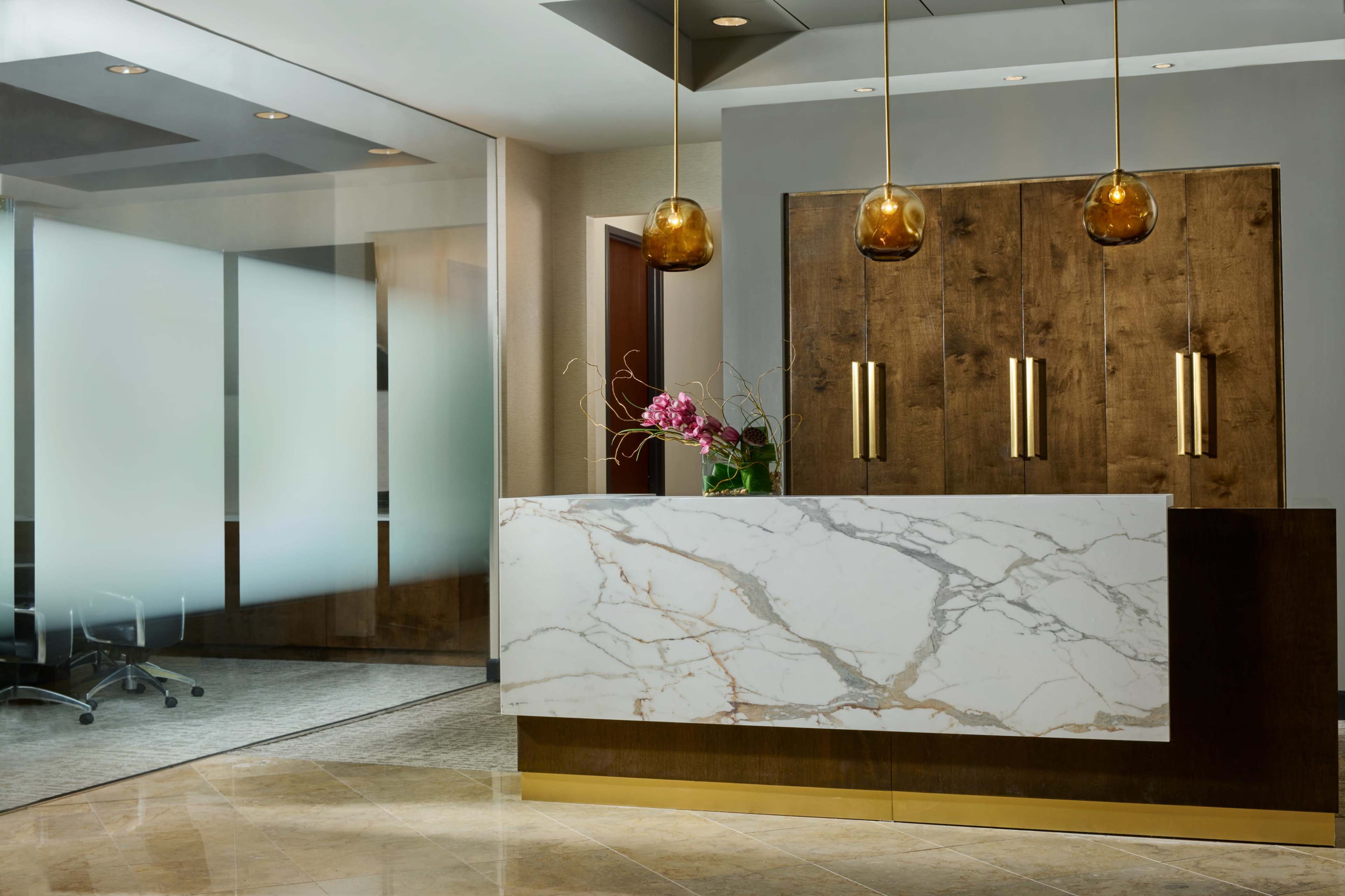 A modern reception area features a marble front desk with gold accents and wooden cabinetry, illuminated by three pendant lights.