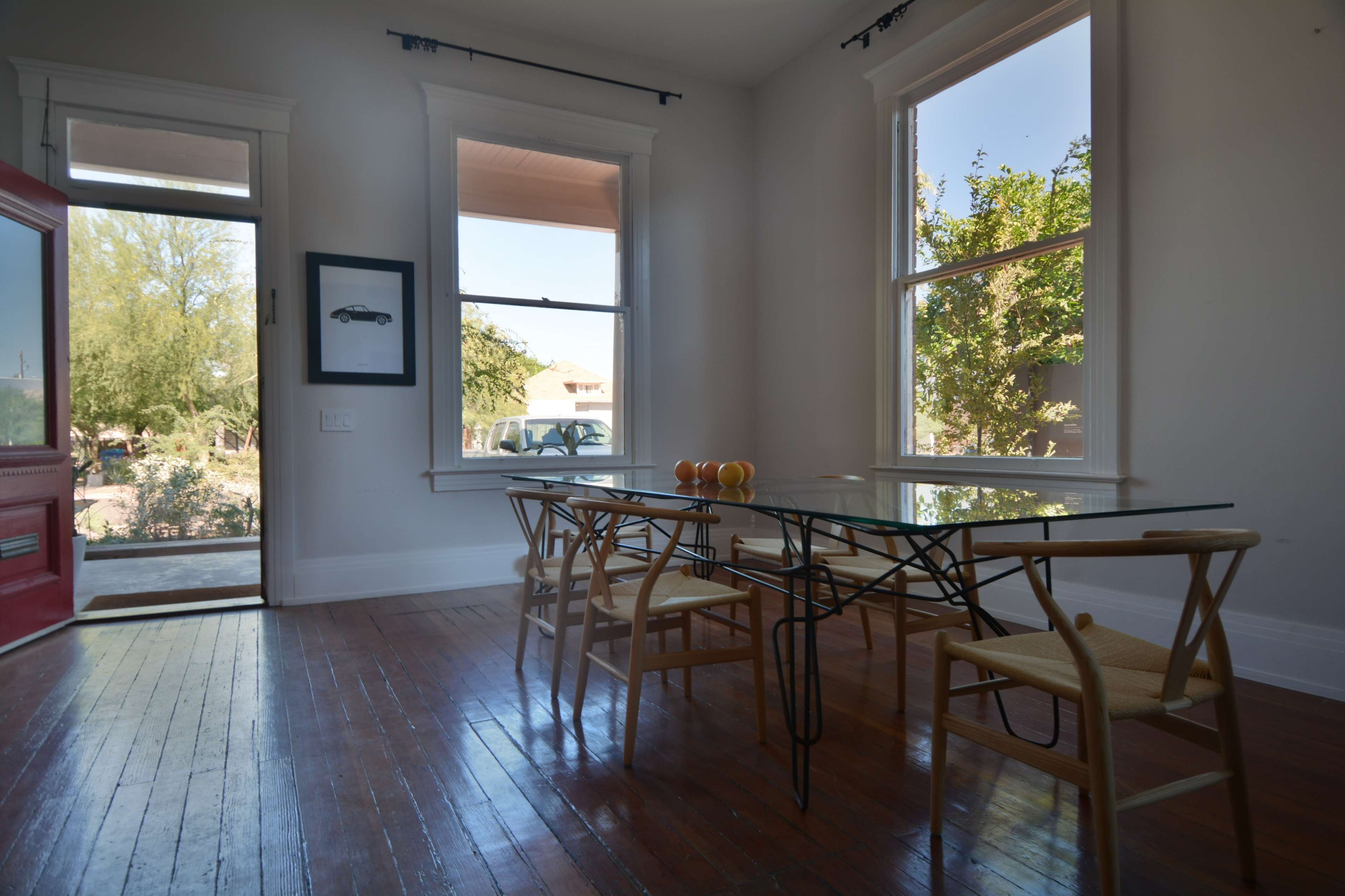 A dining area features a glass table with four wooden chairs, large windows allowing natural light, and a red front door visible in the background.
