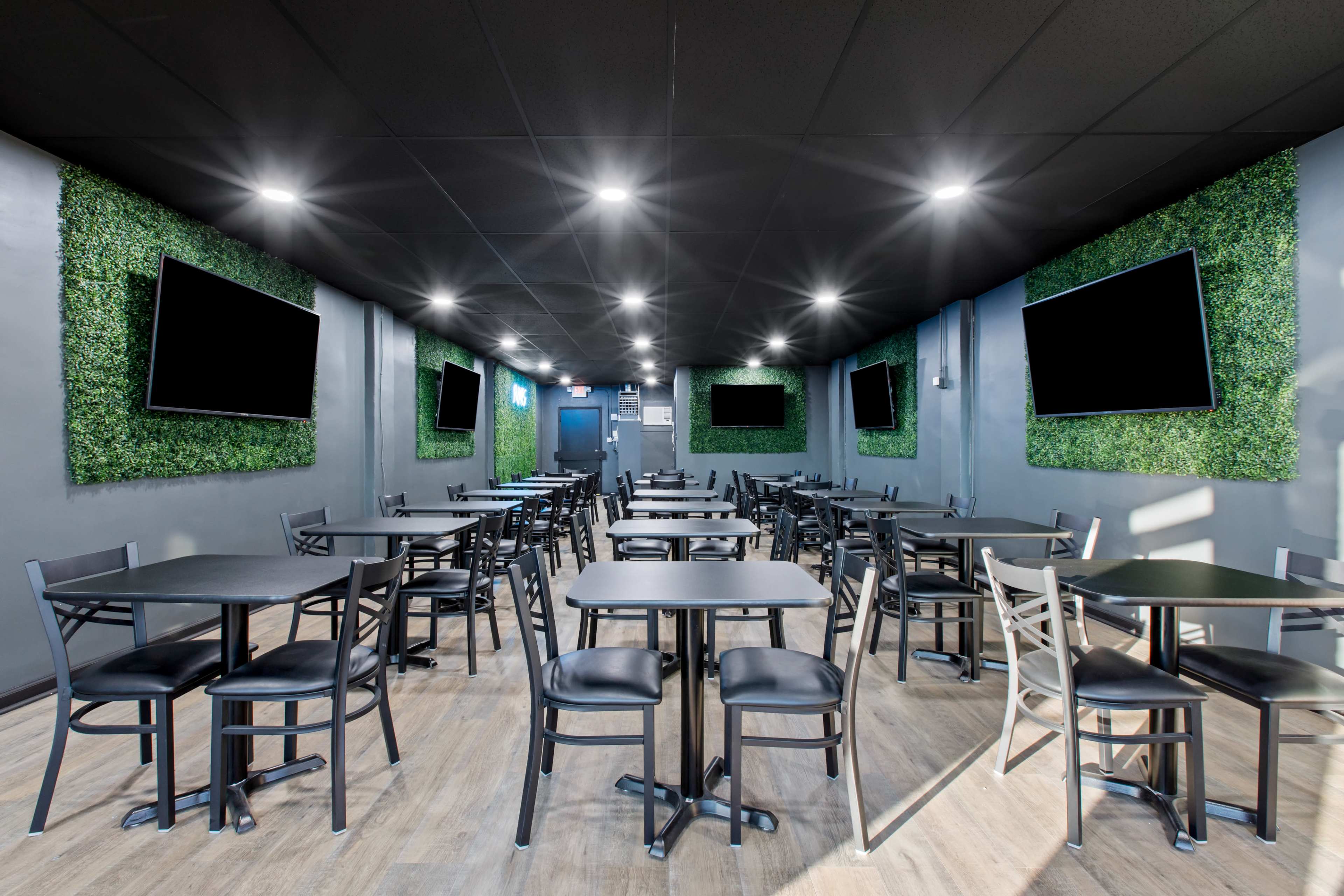 A modern dining area with multiple black tables and chairs, surrounded by green wall panels and equipped with wall-mounted televisions.