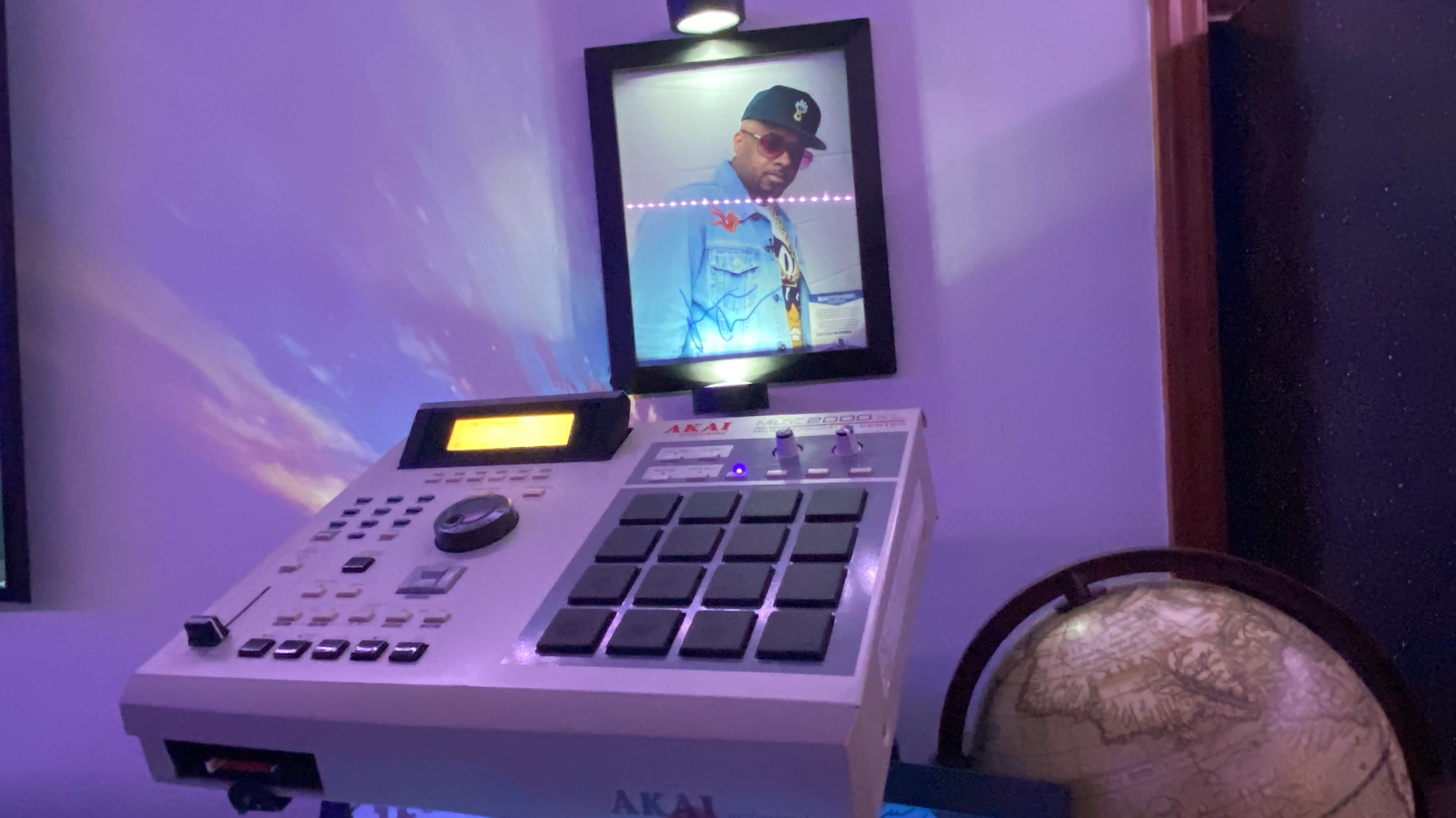 A white AKAI music production controller is positioned on a table under a framed portrait of a man, with a globe visible on the side.