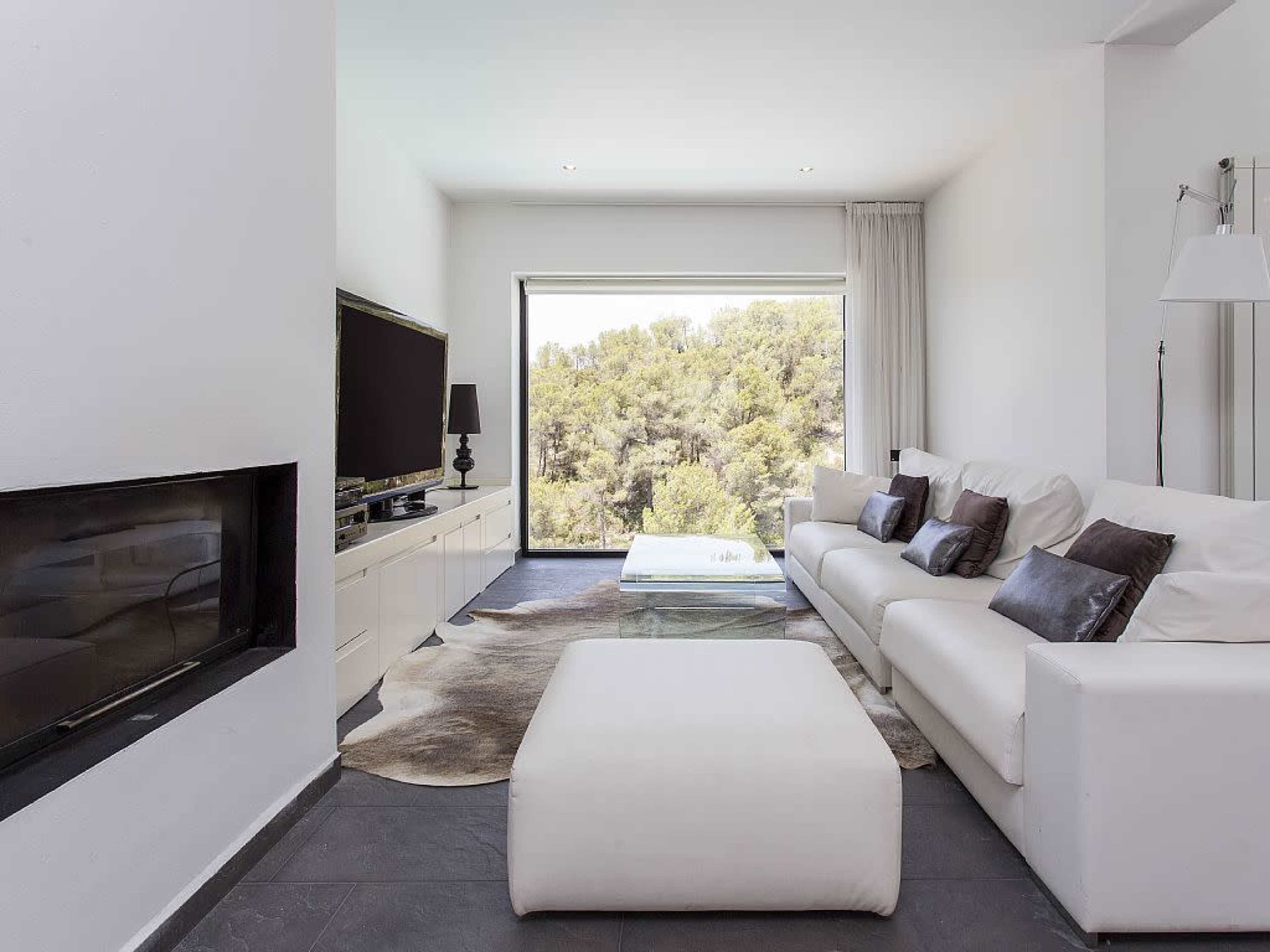 A modern living room featuring a white sofa, a glass coffee table, and a wall-mounted television, with a large window offering a view of greenery outside.