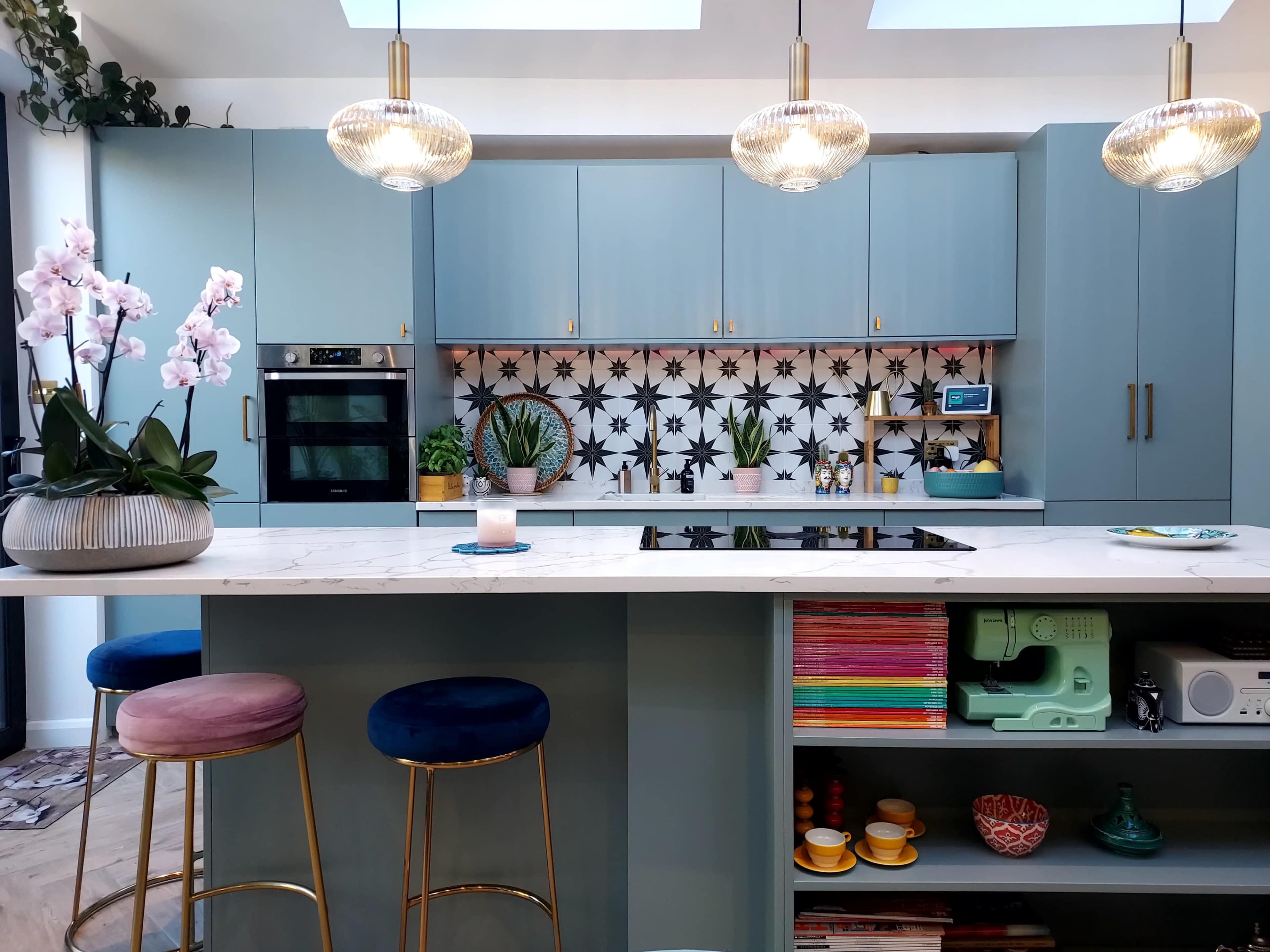 A modern kitchen features blue cabinets, a patterned backsplash, and a central island with bar stools and decorative plants.
