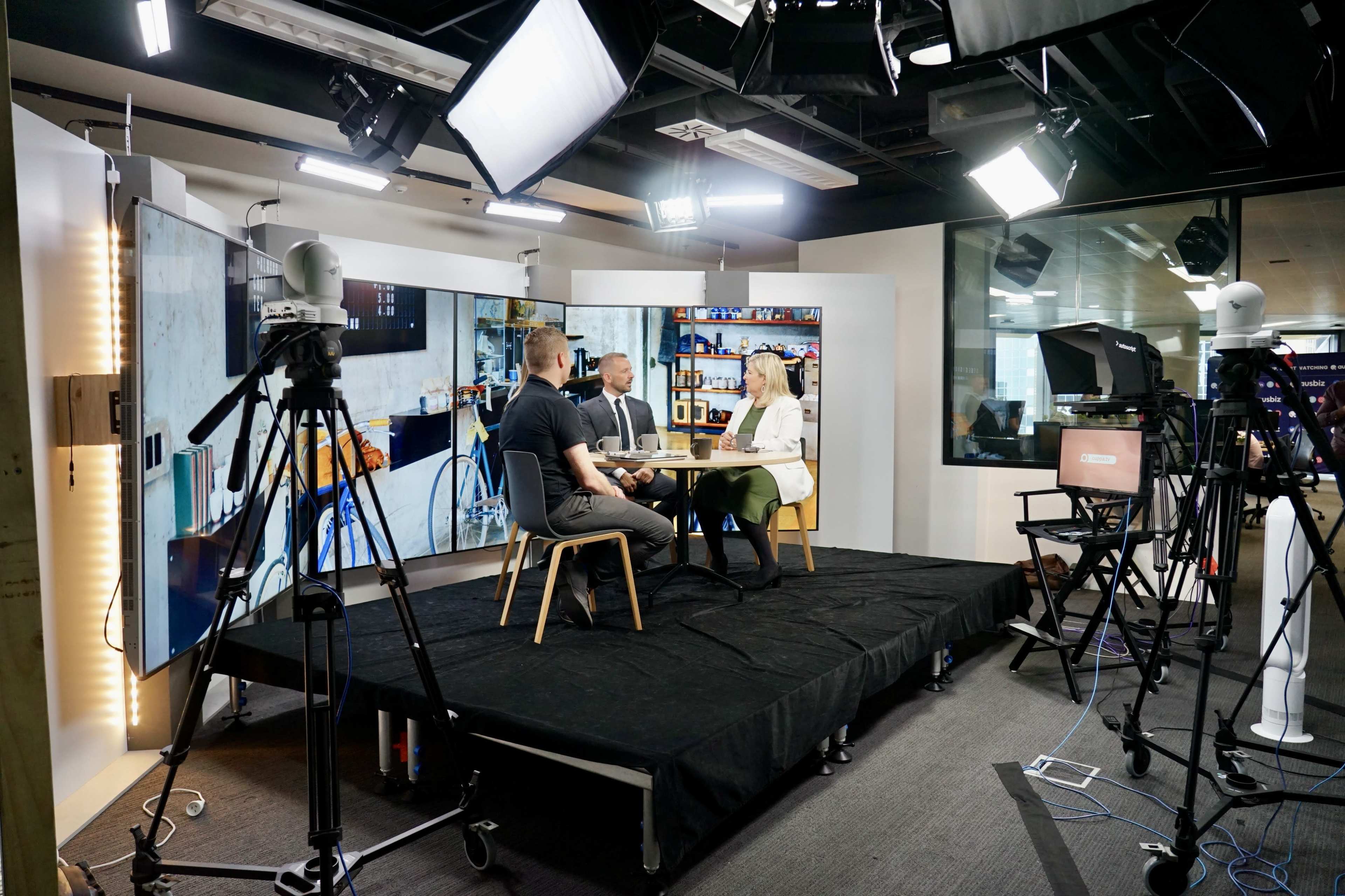 A television studio is set up with three people sitting at a table, surrounded by cameras and studio lights.