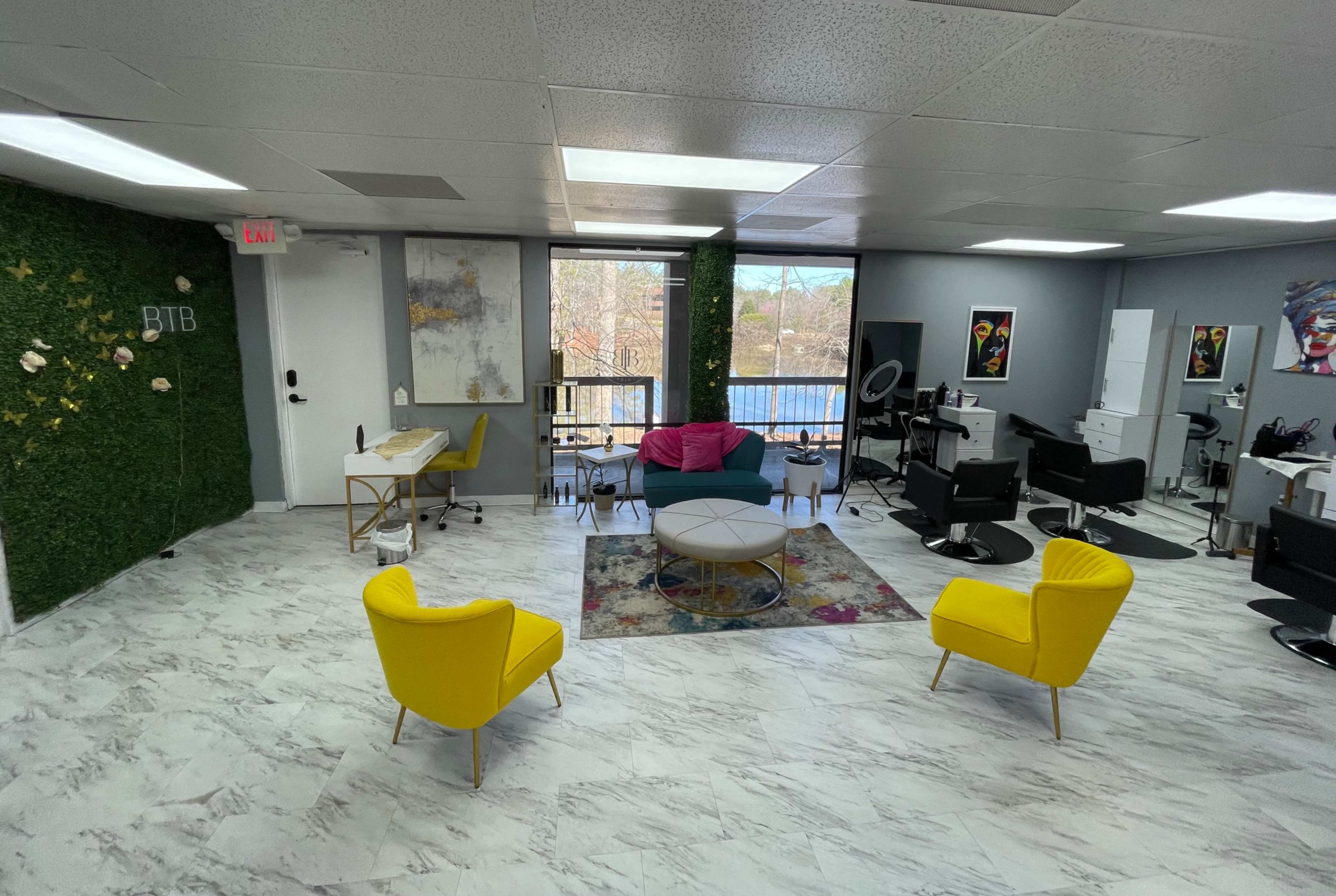 A modern salon interior with yellow chairs, a circular coffee table, a large window overlooking a body of water, and multiple styling stations.