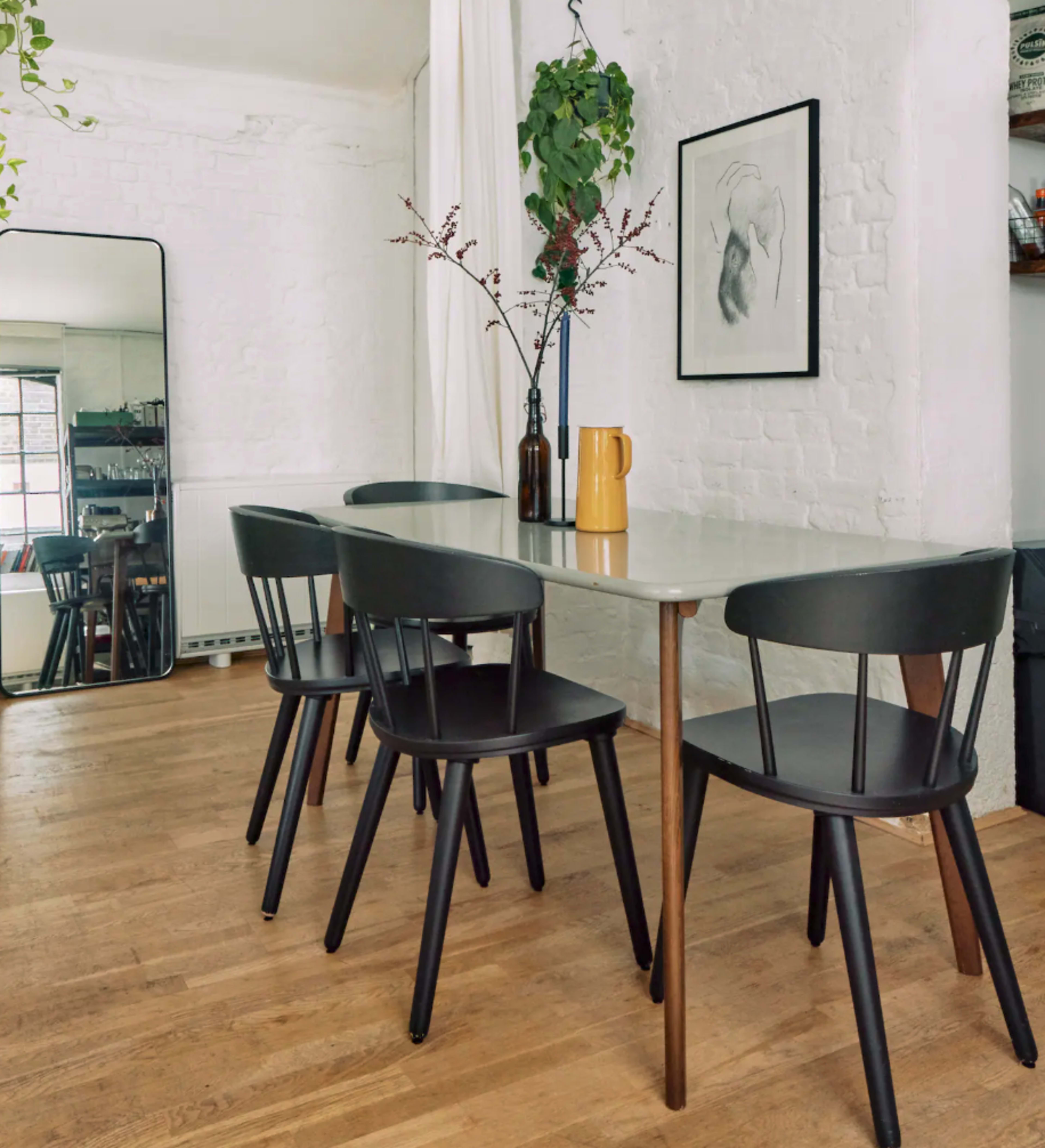 A minimalist dining area features a long table with four black chairs, a mirror, and a potted plant, set against a white brick wall.