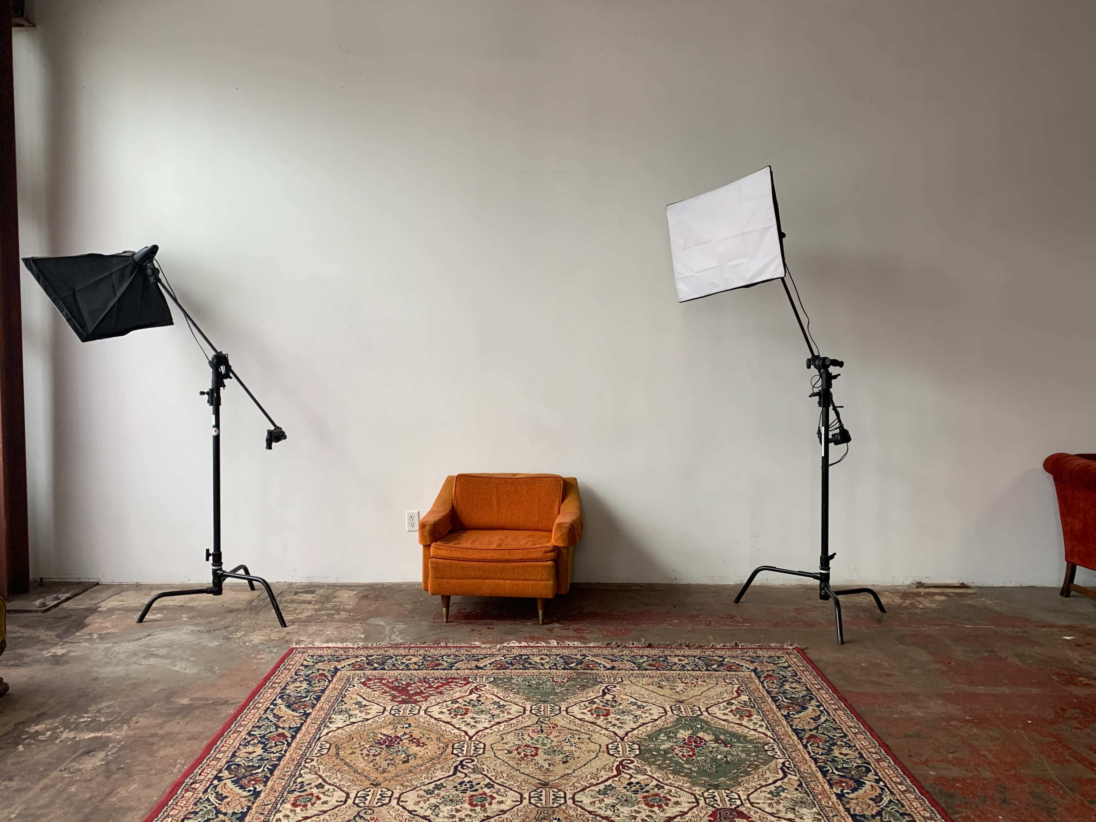 A small orange chair is positioned on a patterned rug between two studio lights in an empty room.