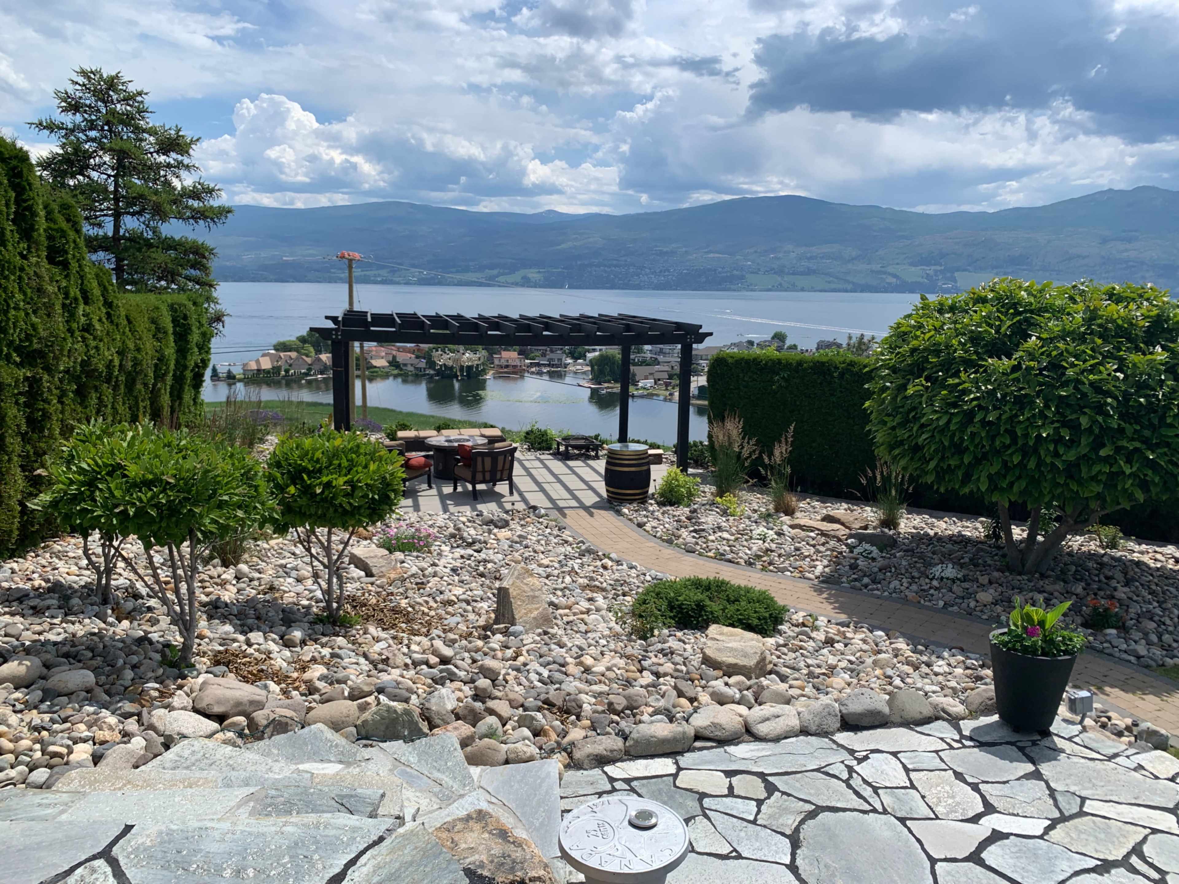 A landscaped garden with a stone pathway leading to a seating area under a pergola, overlooking a lake and mountains.