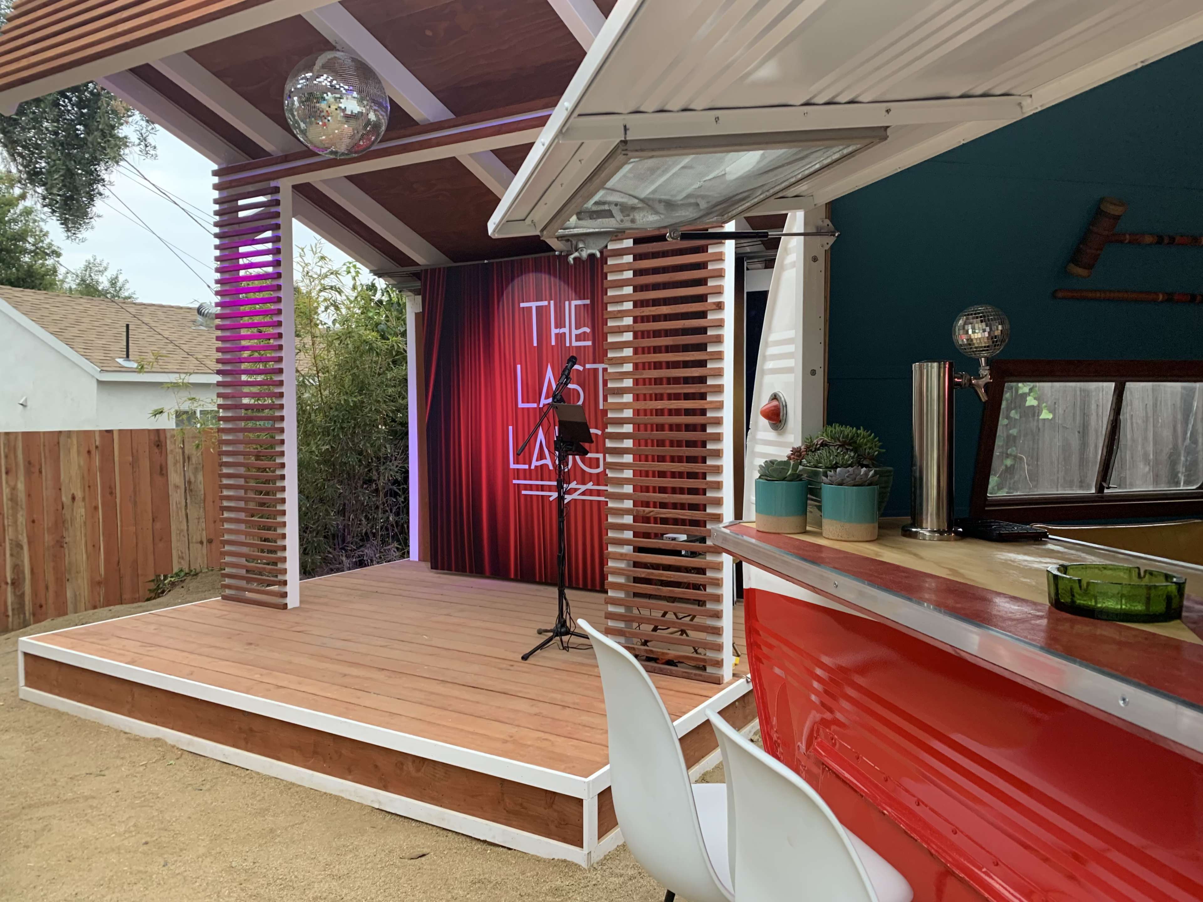 A small outdoor stage with a microphone in front of a red backdrop featuring the words "THE LAST LAUGH," and a nearby bar area with white stools and potted plants.