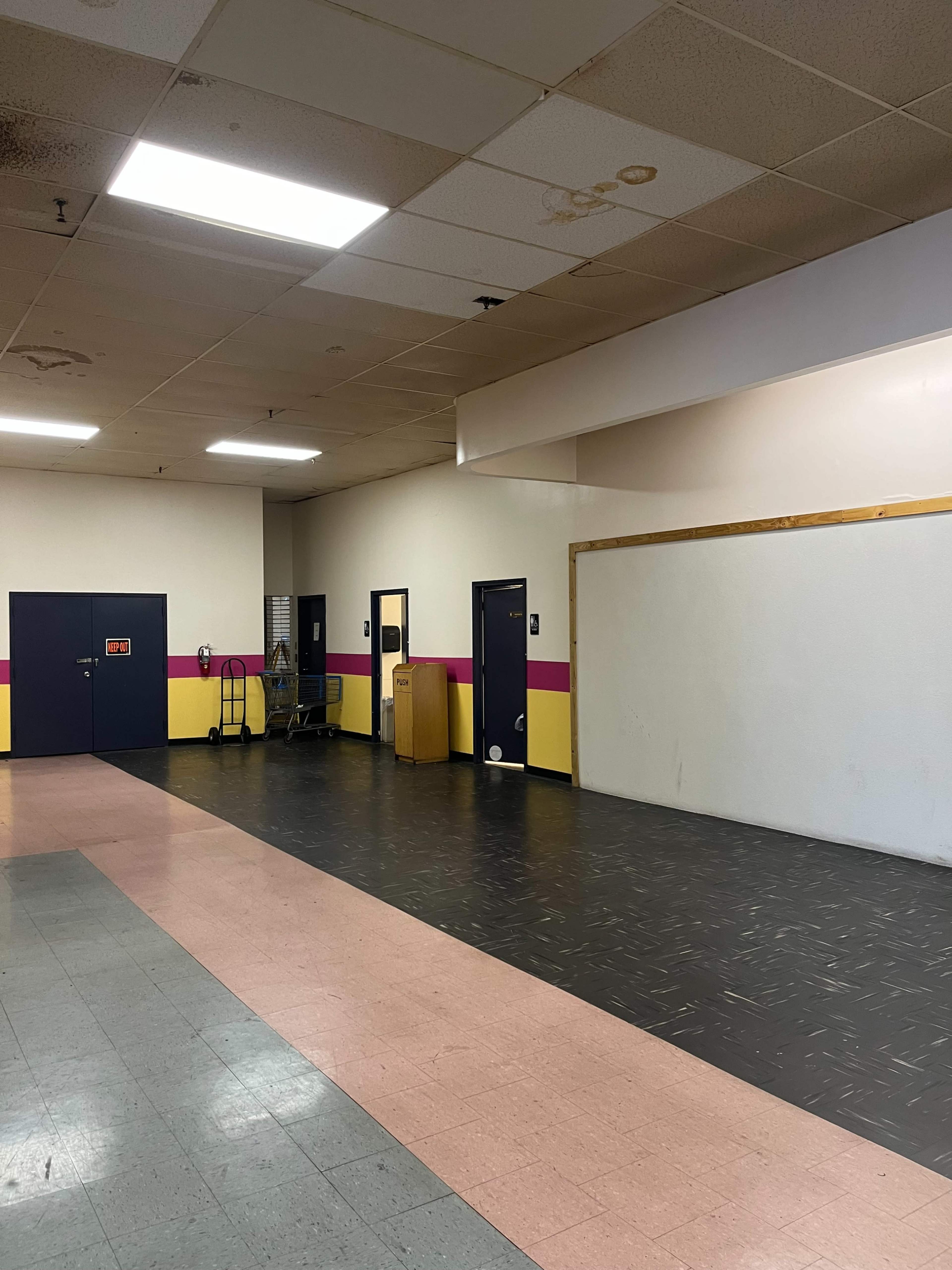 A sparsely furnished indoor hallway with a dark floor and walls painted in pink and yellow, leading to several doors.