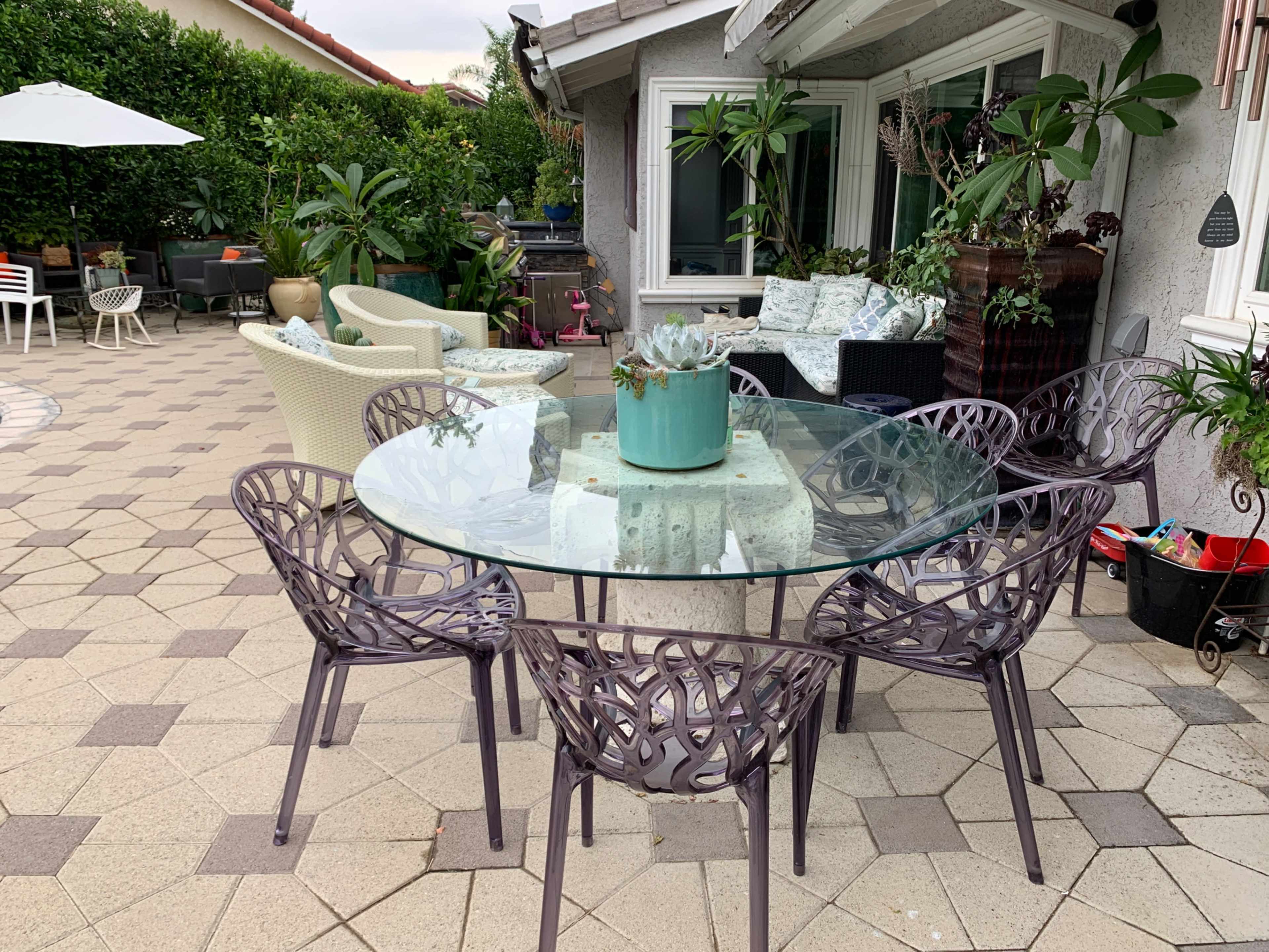 A circular glass table surrounded by six patterned chairs on a tiled patio, with various potted plants and outdoor furniture in the background.