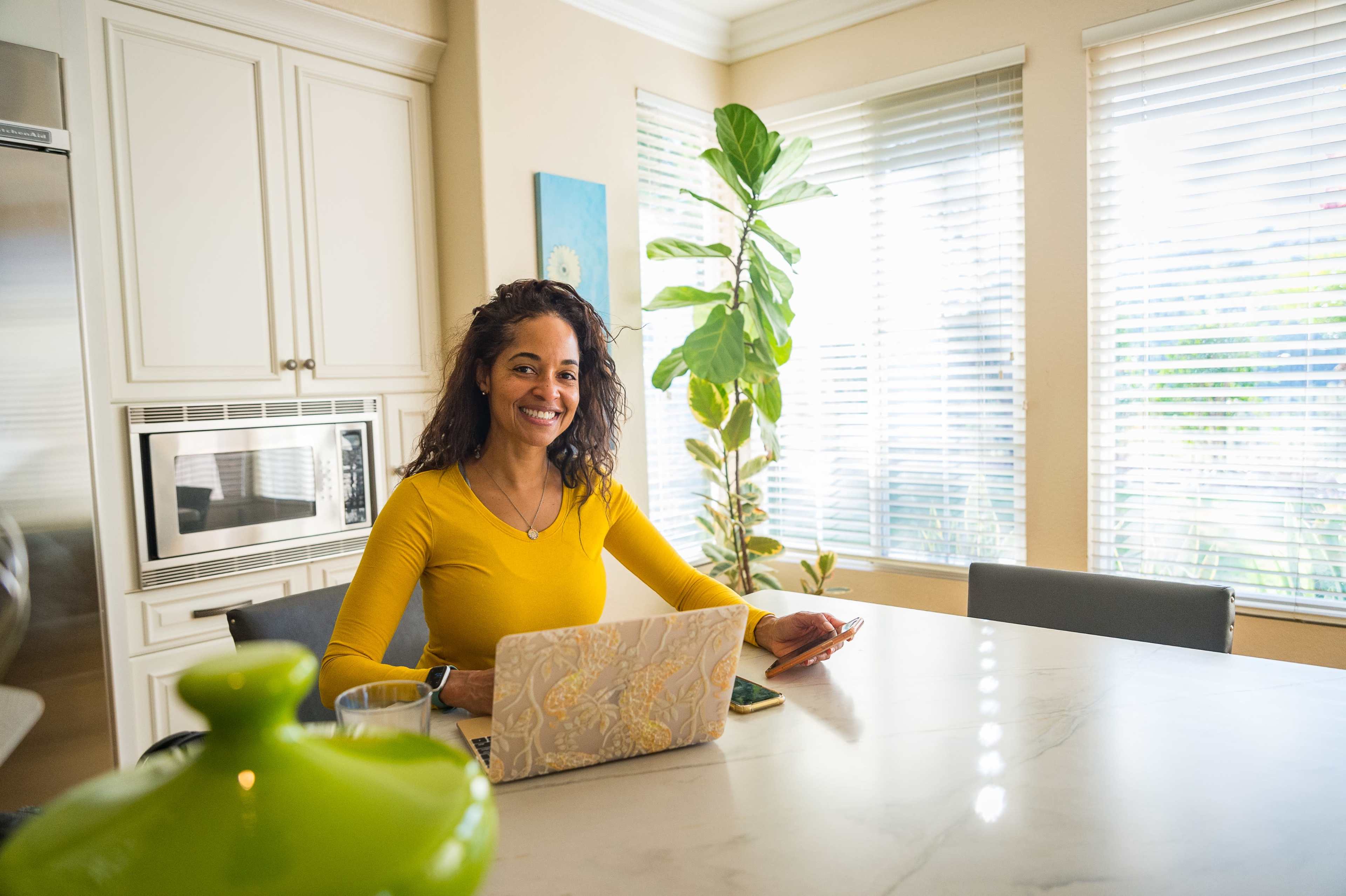 A woman with curly hair sits at a kitchen table with a laptop and a smartphone, in a well-lit room adorned with a potted plant.