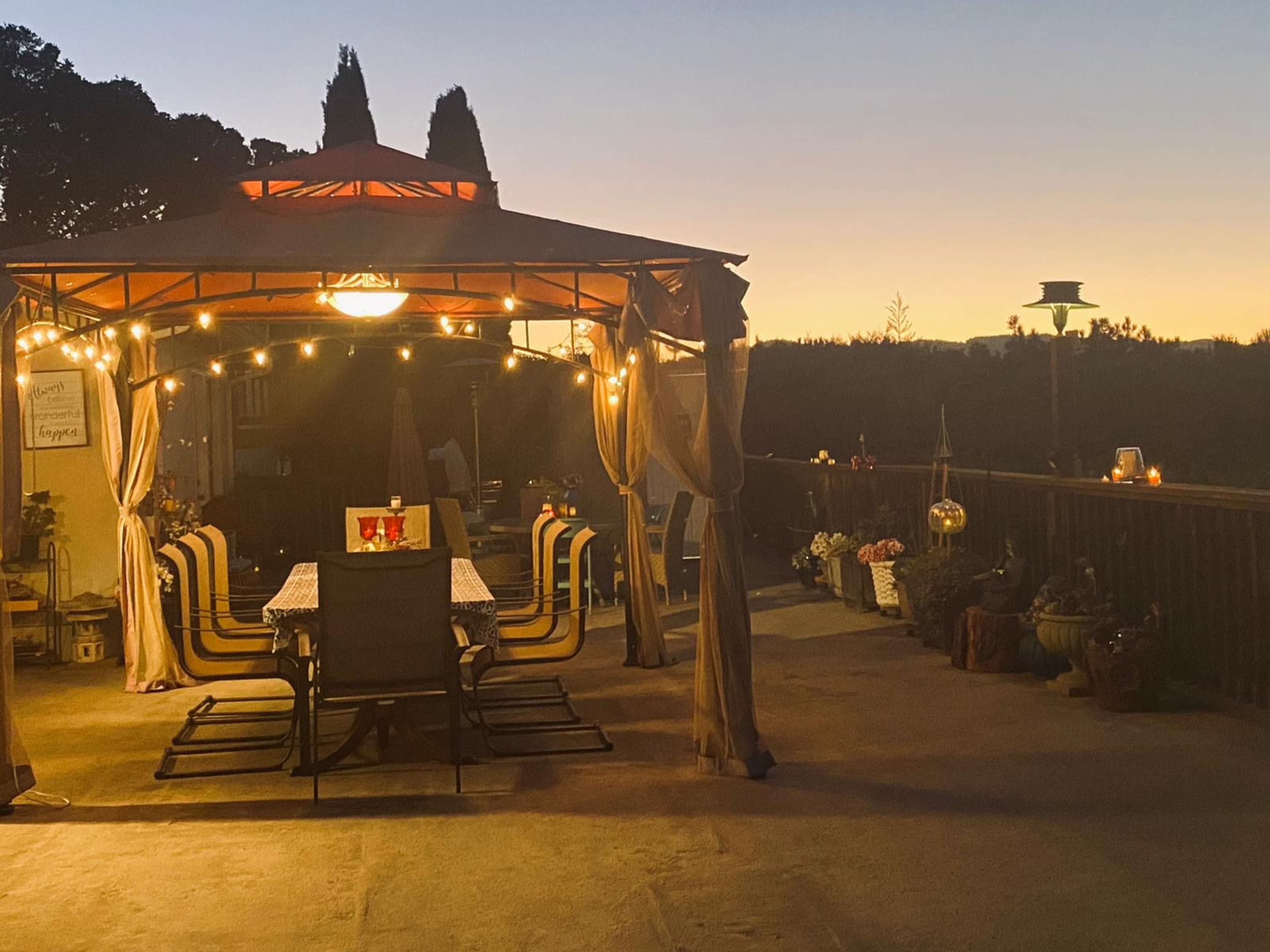 A gazebo with a dining setup and string lights is seen on a patio at sunset, with decorative plants surrounding the area.