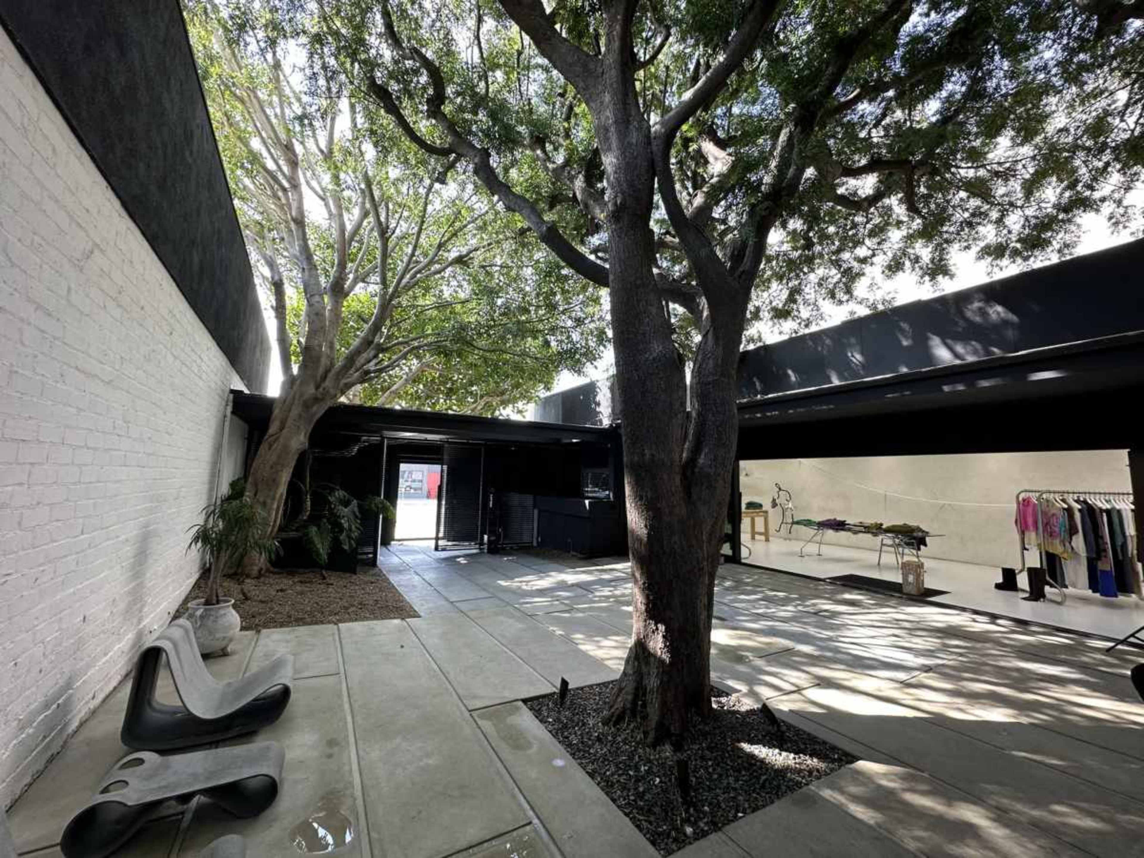 The image shows a spacious outdoor area featuring two large trees, a paved floor, and clothing displays along one wall.
