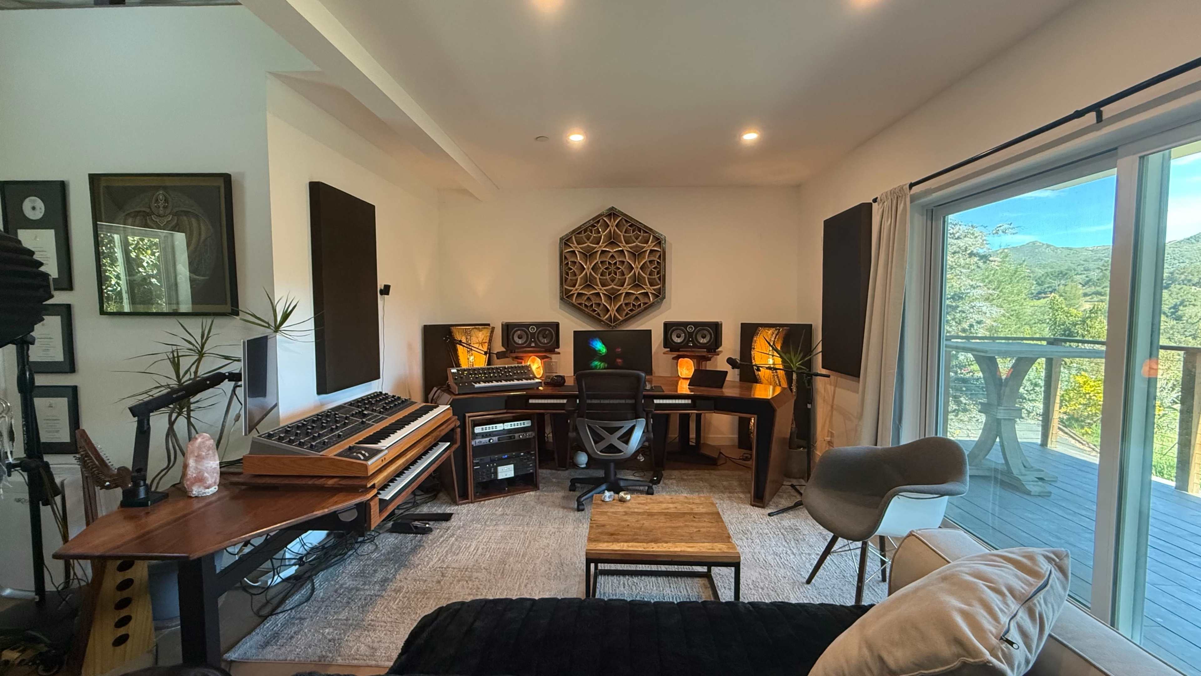 A modern music studio features a desk with audio equipment, a synthesizer on the side, and large windows overlooking a natural landscape.