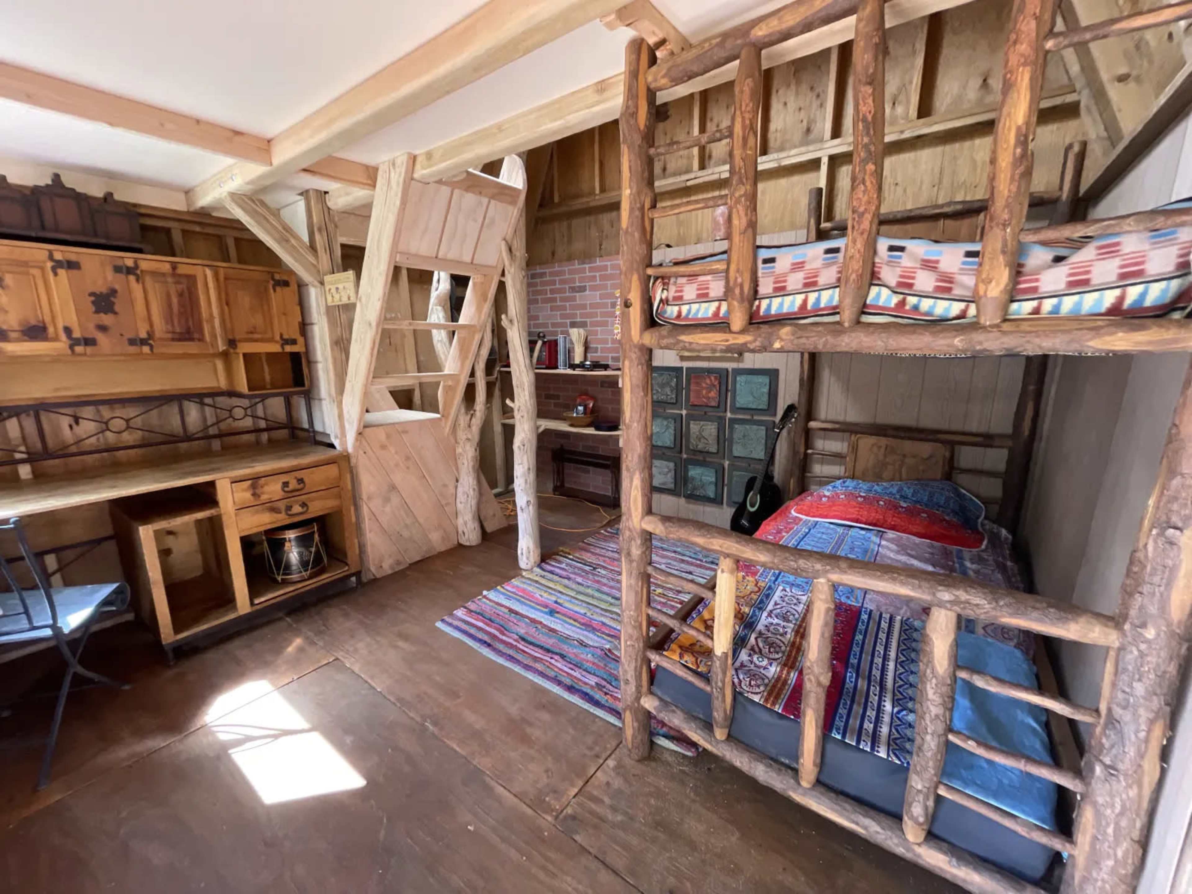 The image shows a rustic bedroom with a wooden loft bed, a lower sleeping area, a desk, and colorful textiles on the floor and beds.