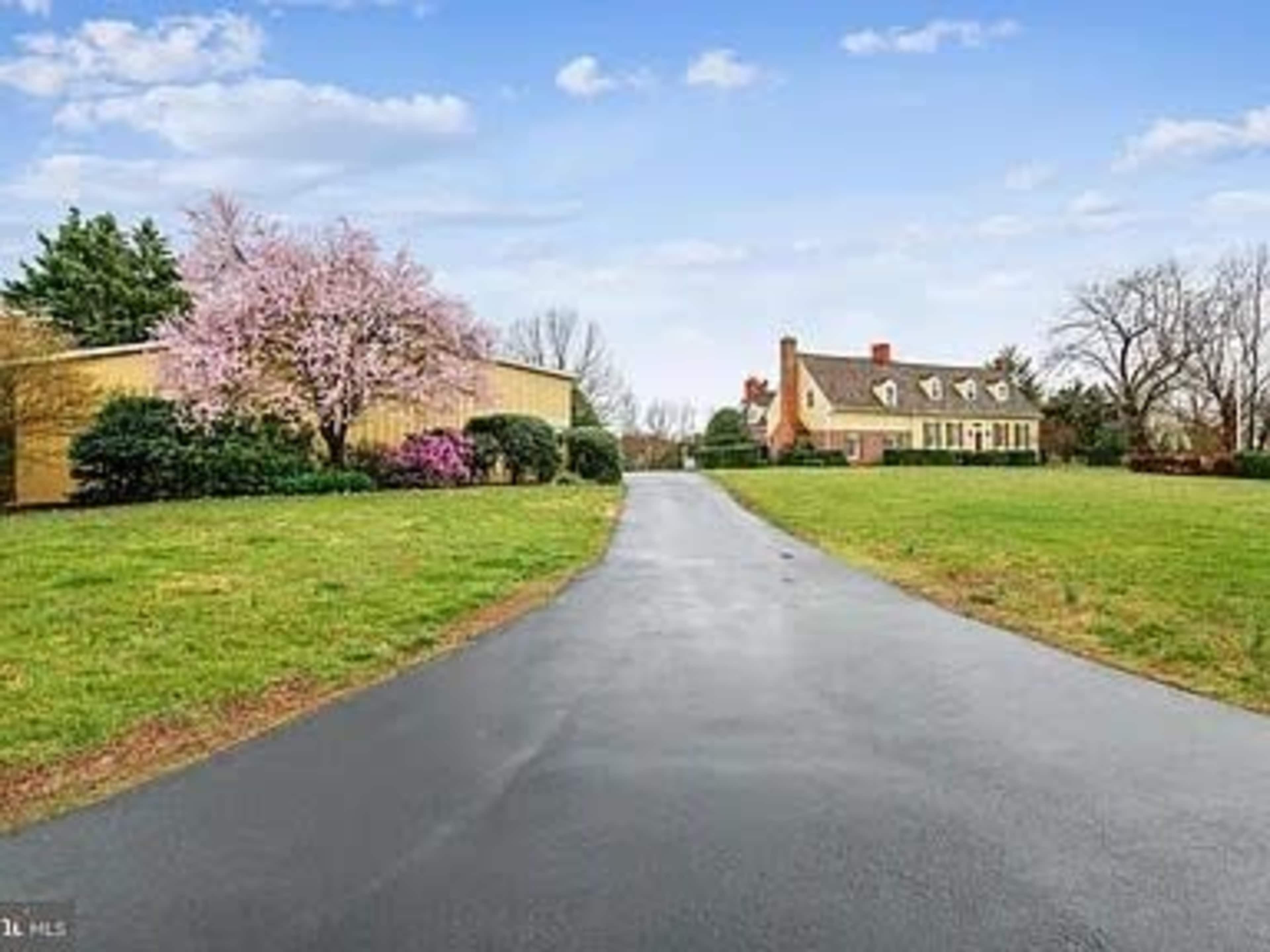 A paved driveway leads to a large house surrounded by greenery and blooming trees.