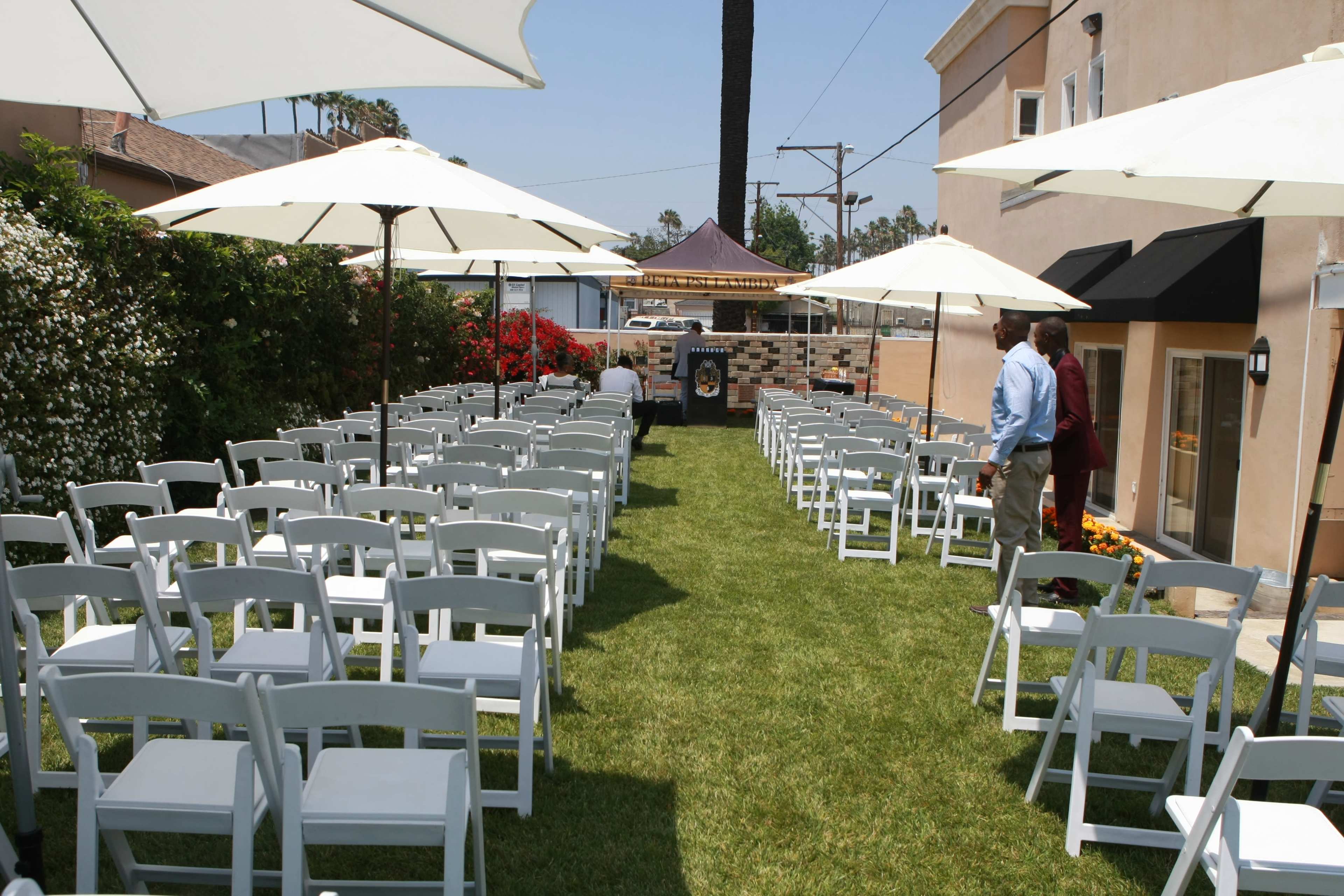 A grassy outdoor space is set up with white chairs arranged in rows under large umbrellas, leading to a small stage at the far end.