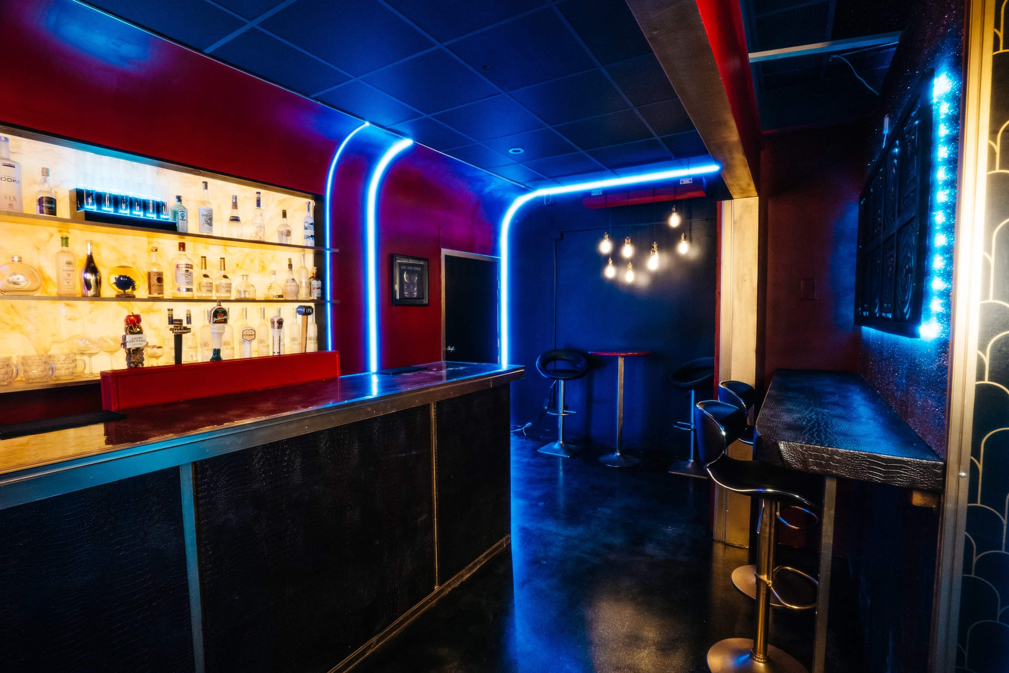 A modern bar area with a brightly lit backlit shelf displaying various bottles and seating along the walls.