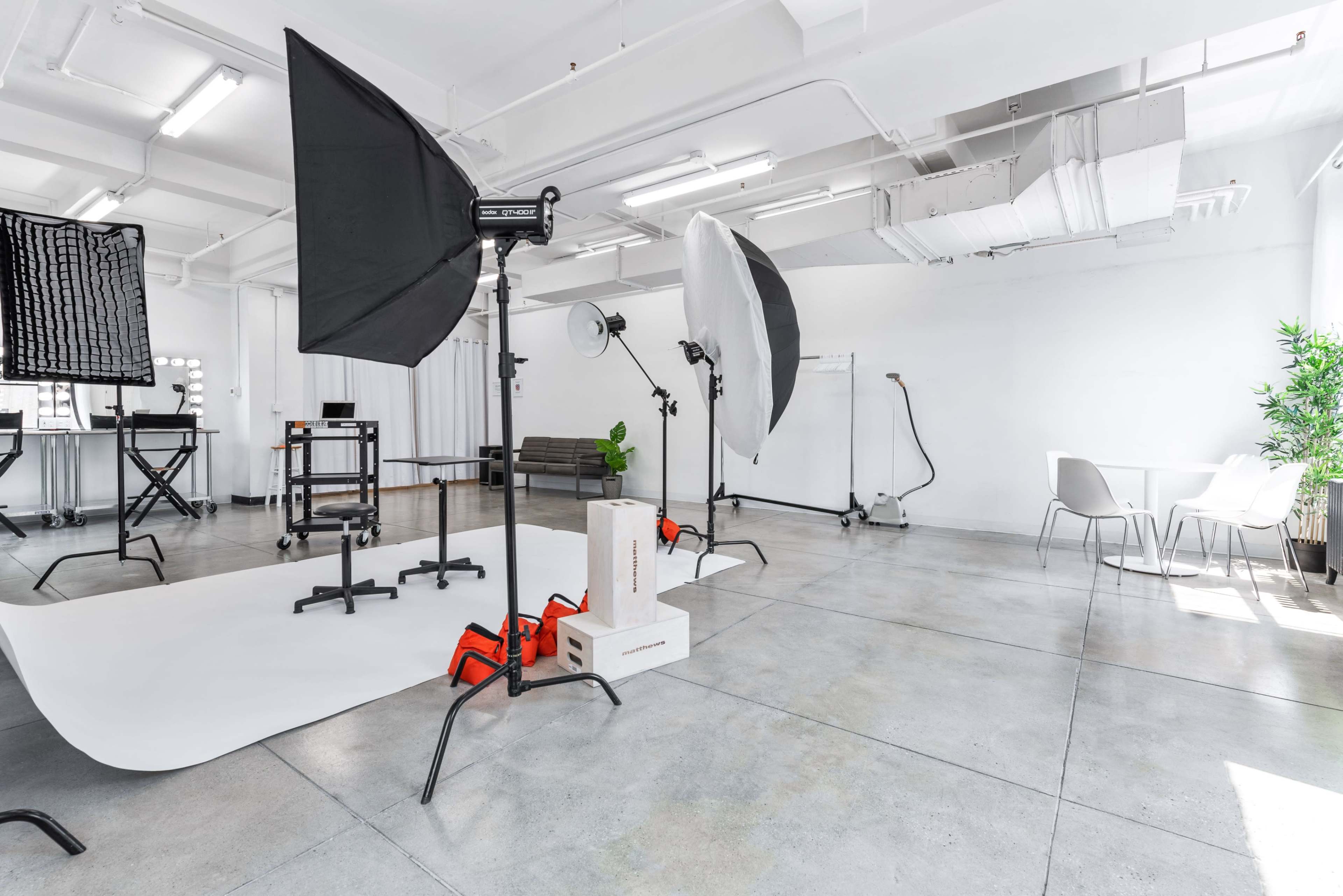 A spacious, well-lit photo studio features various lighting equipment, a white backdrop, and minimalist furniture.