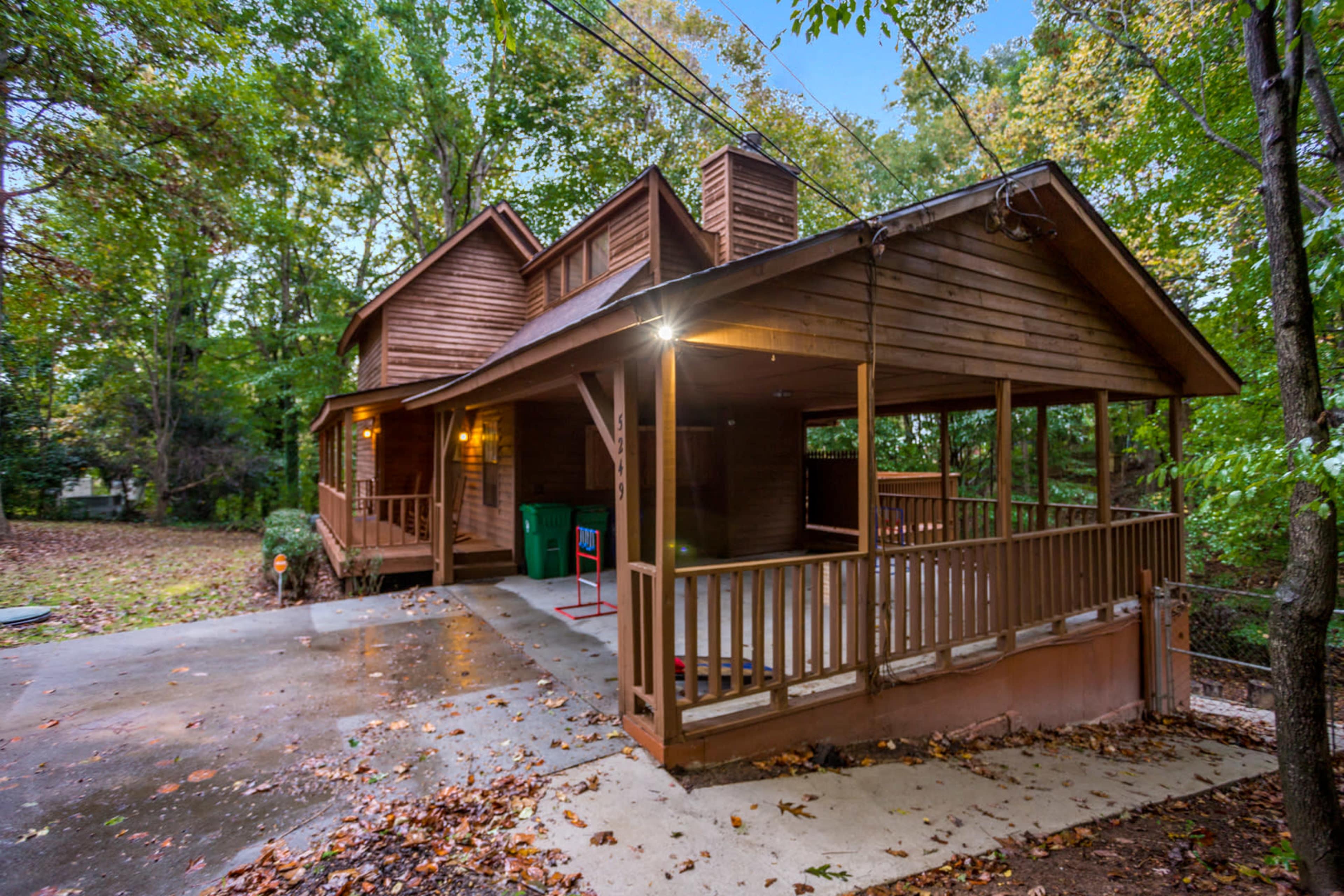 A wooden house with a porch, surrounded by trees, sits on a paved area with scattered leaves.